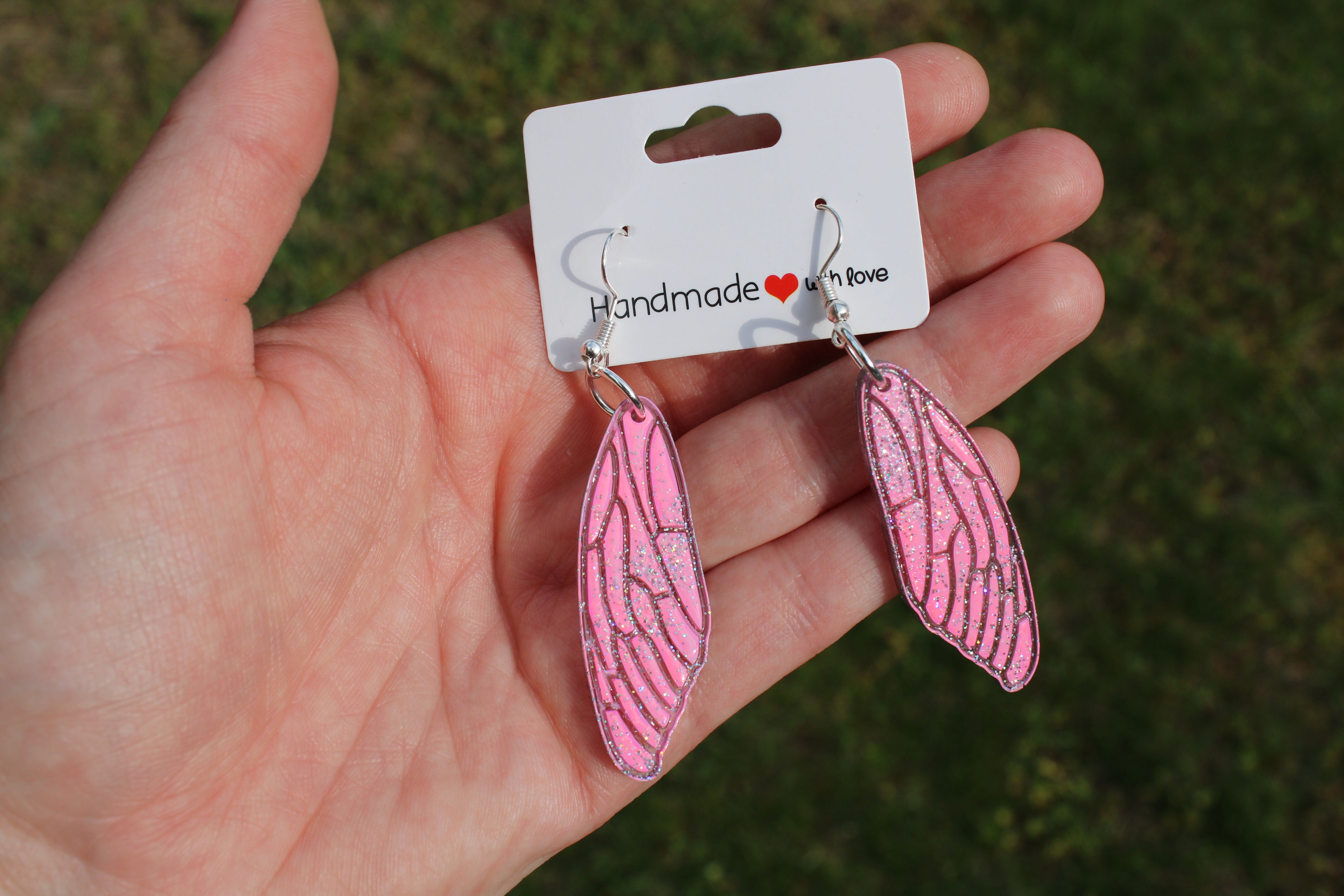 Handmade pink wing-shaped earrings on a card held in a hand with a grassy background