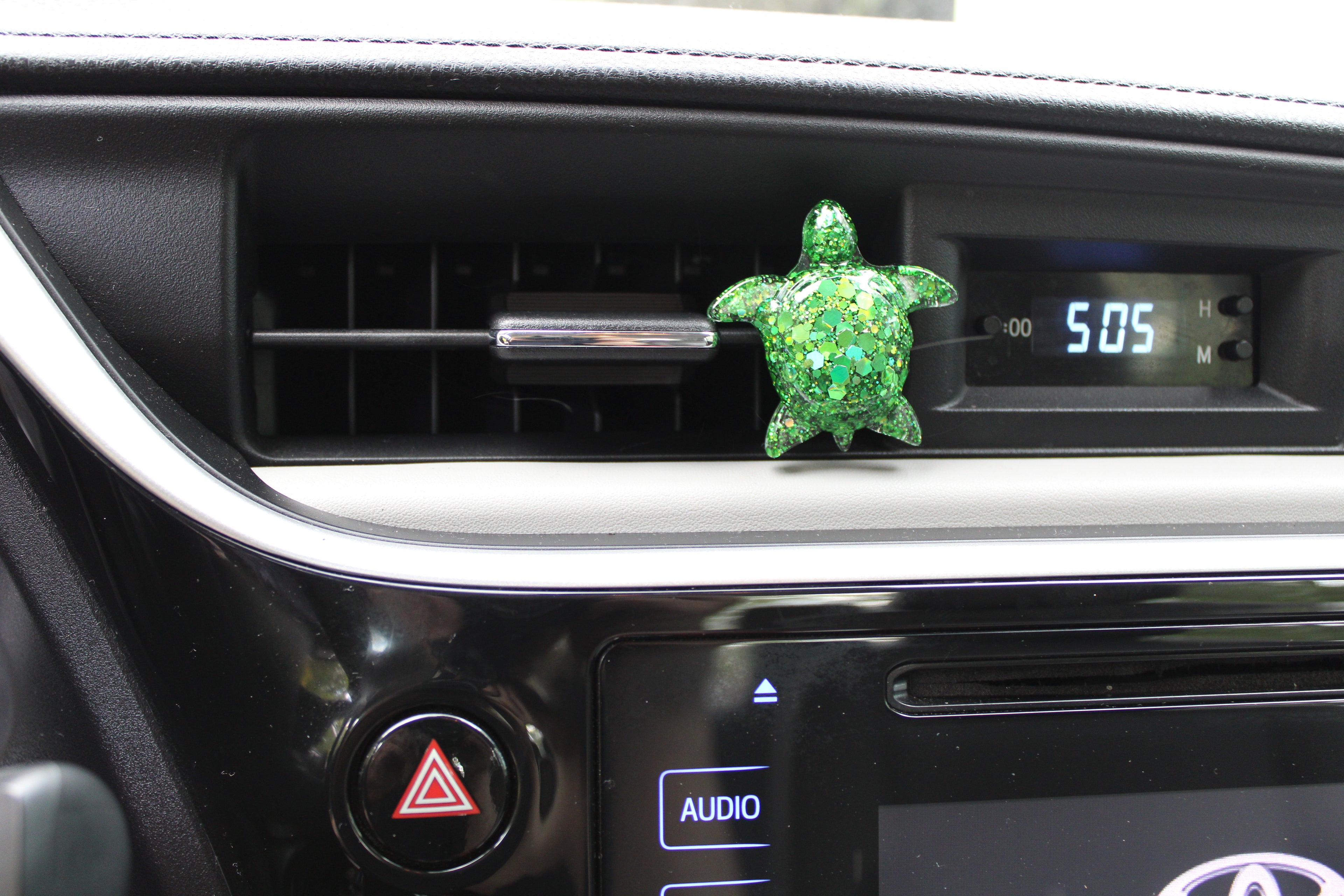 Green turtle-shaped car vent clip on a car dashboard with digital clock display.
