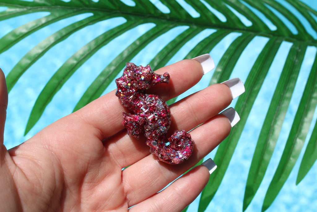 Hand holding a glittery pink seahorse-shaped car vent clip against a palm leaf background