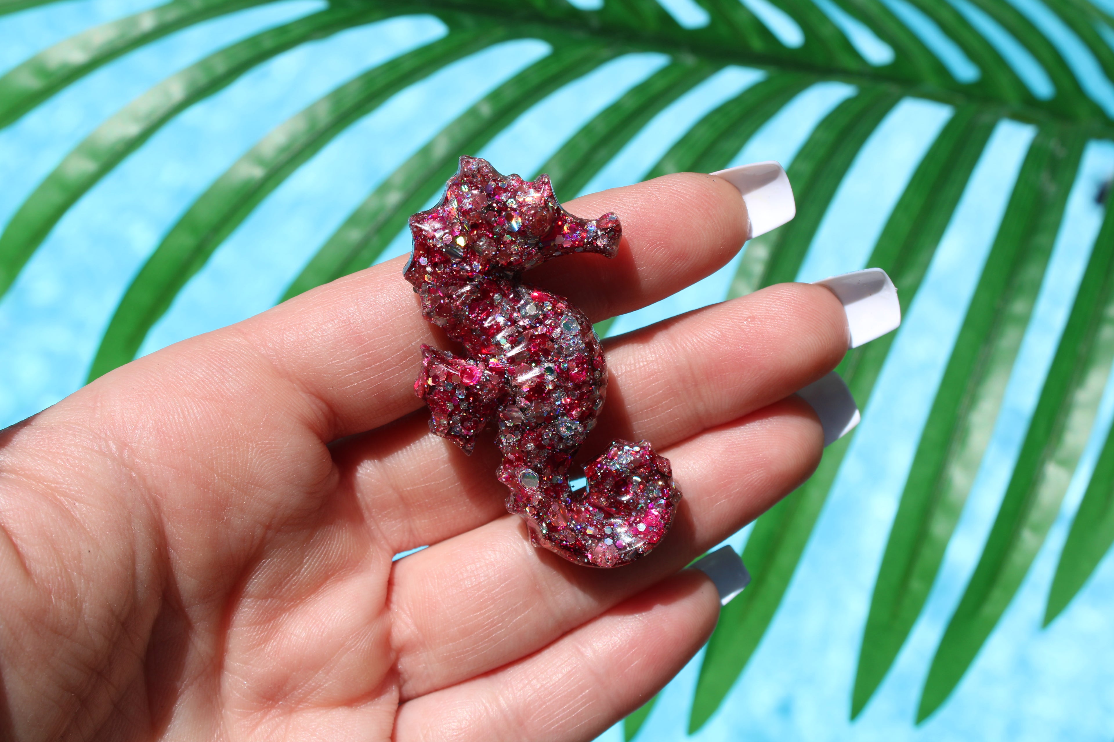 Hand holding a pink glittery seahorse car vent clip against a palm leaf background