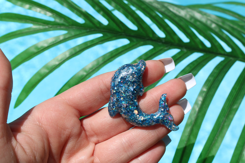 Hand holding a glittery blue dolphin car vent clip with palm leaves in the background