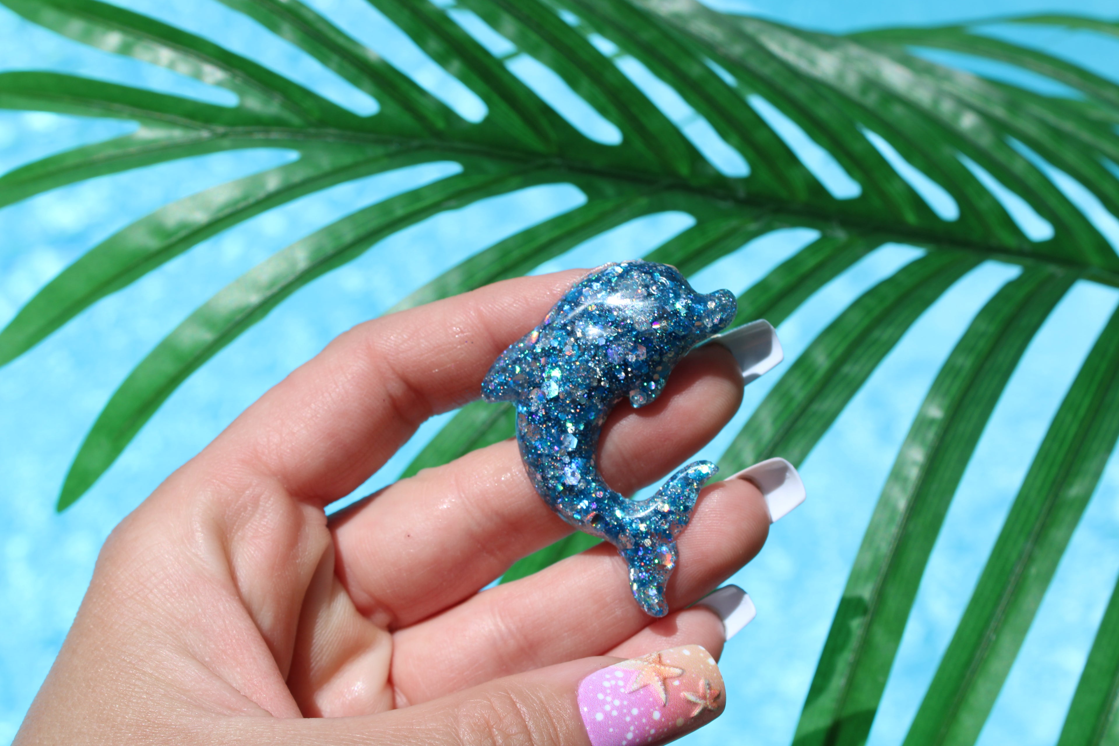 Hand holding a glittery blue dolphin vent clip with palm leaves in the background