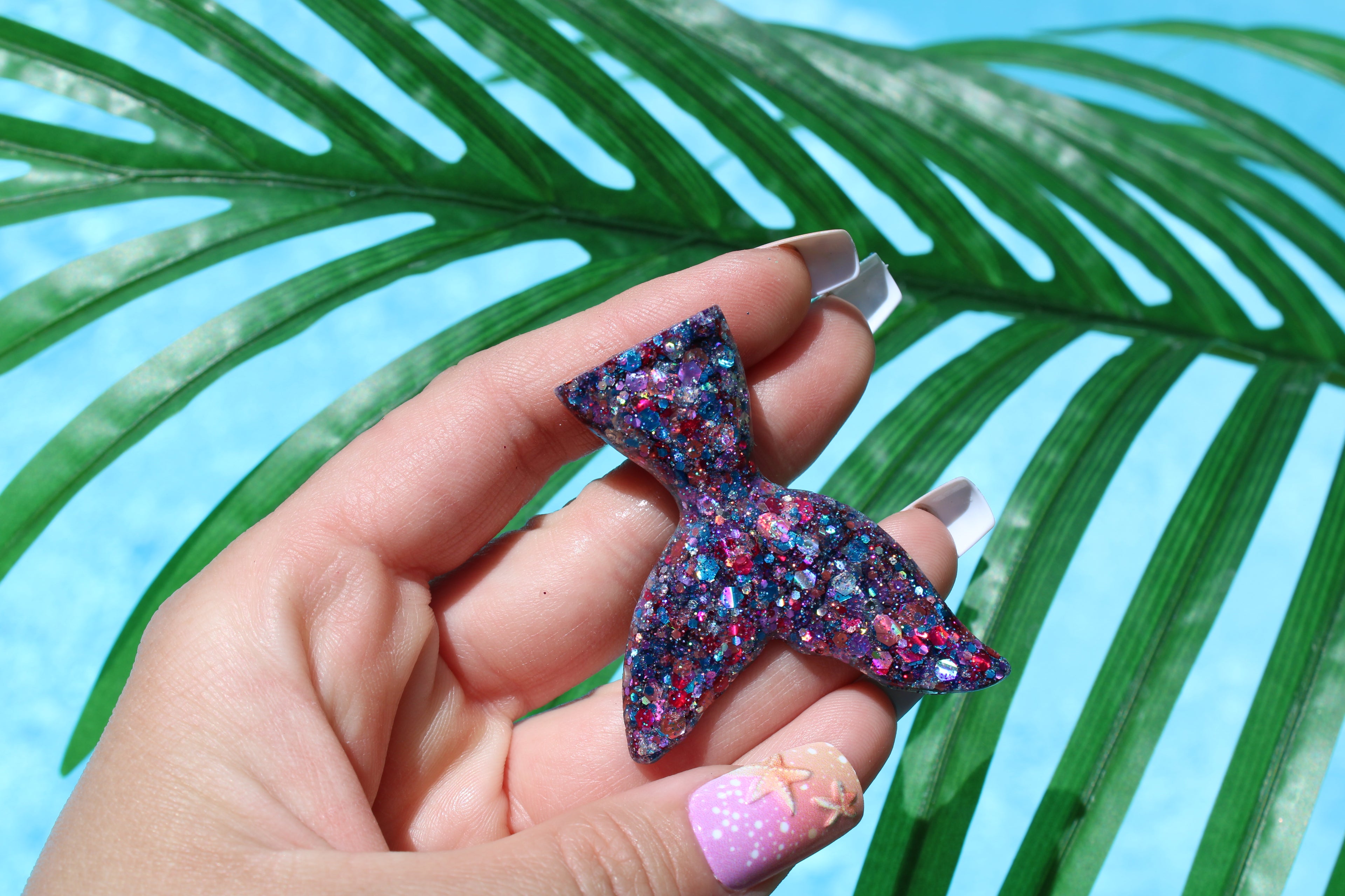 Hand holding a glittery mermaid tail vent clip with palm leaves in the background