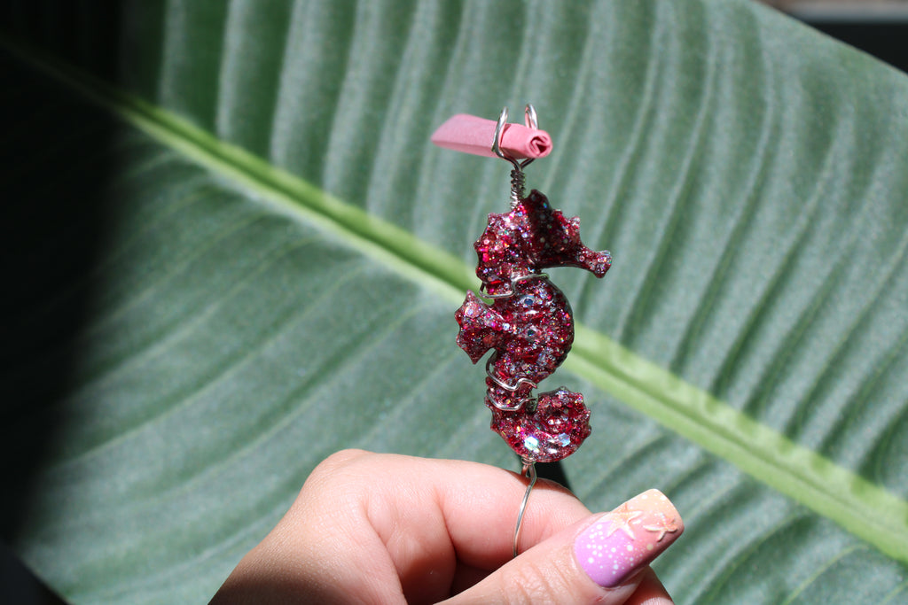 Hand holding a pink sequin resin seahorse smoke ring in front of a leaf