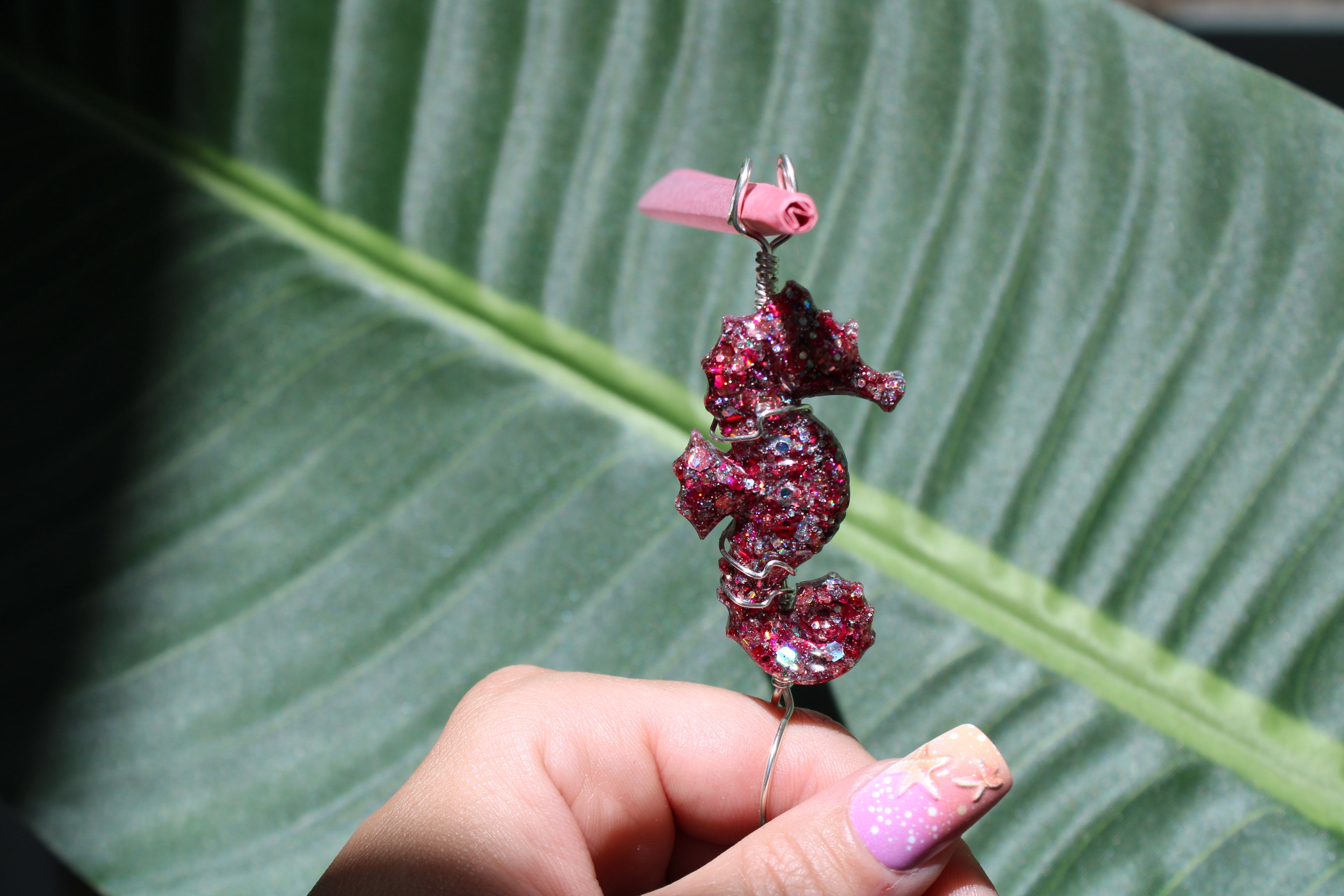 Hand holding a pink sequin resin seahorse smoke ring in front of a leaf