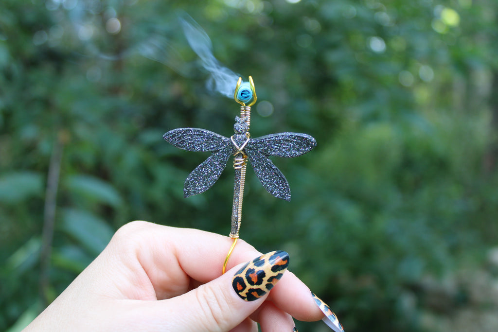 A hand with leopard-print nail art holds a dragonfly-shaped smoke ring with a black glitter body, gold wire details against a blurred green outdoor background.