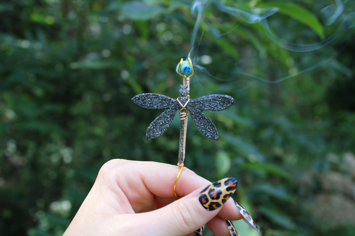 A hand with leopard-print nails holds a smoking dragonfly joint holder against a blurred green outdoor background. The dragonfly has glittery black wings and body with gold wire details.
