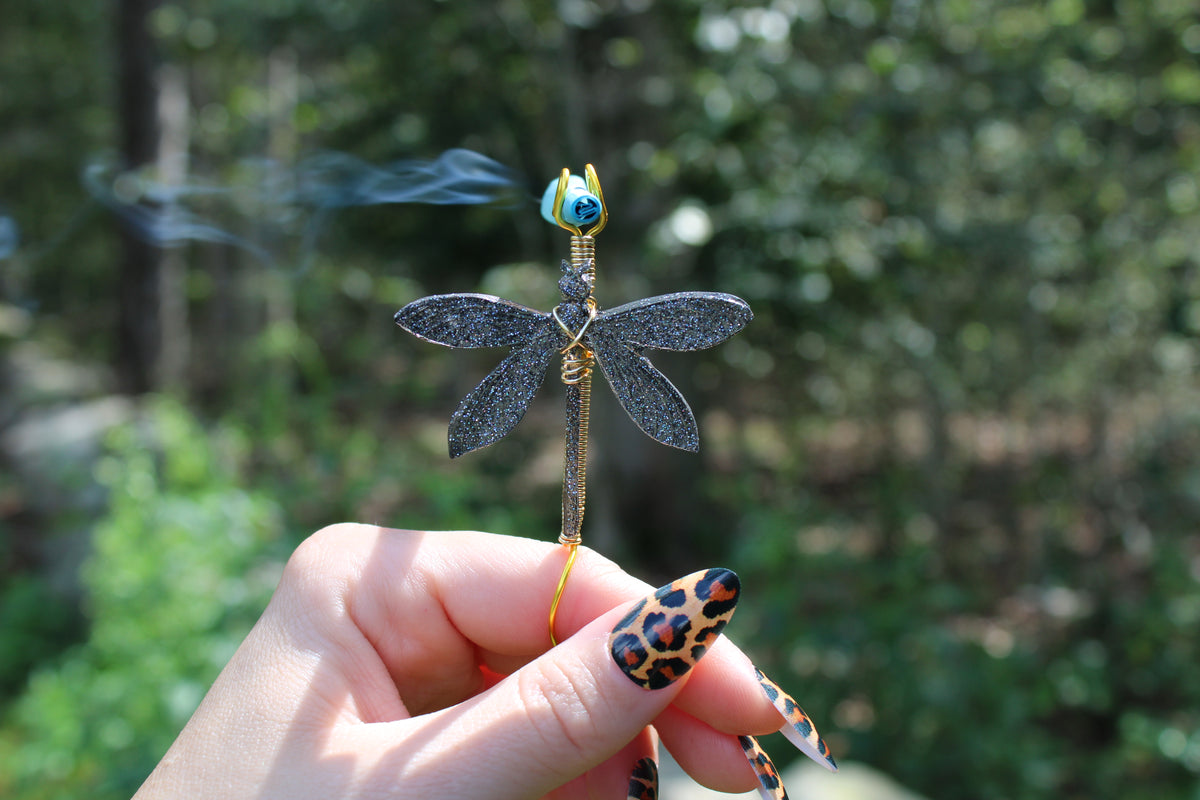 A hand with leopard-print nails holds a smoking dragonfly joint holder outdoors against a blurred green forest background.