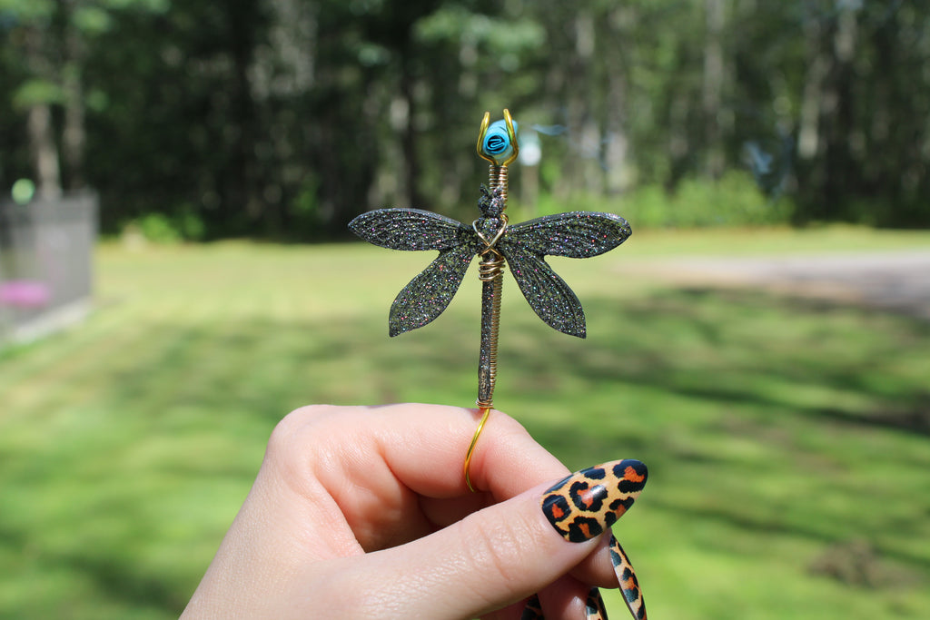 A hand with leopard-print nails holds a metallic dragonfly smoke ring against a blurred outdoor background of green grass and trees.