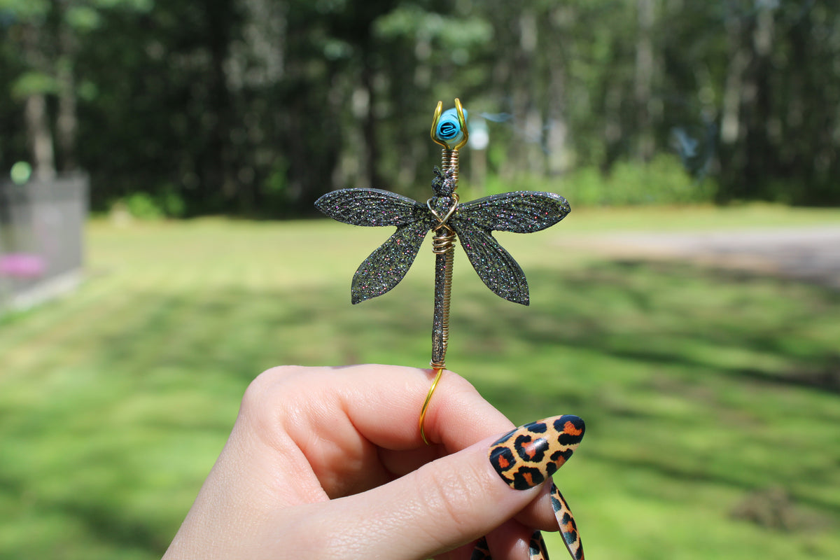 A hand with leopard-print nails holds a metallic dragonfly smoke ring against a blurred outdoor background of green grass and trees.