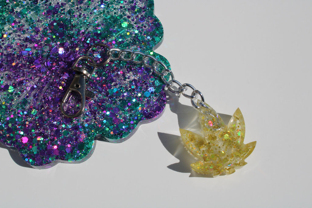 Sparkling resin art features a teal and purple glittery coaster connected to a golden marijuana leaf keychain with a silver chain.