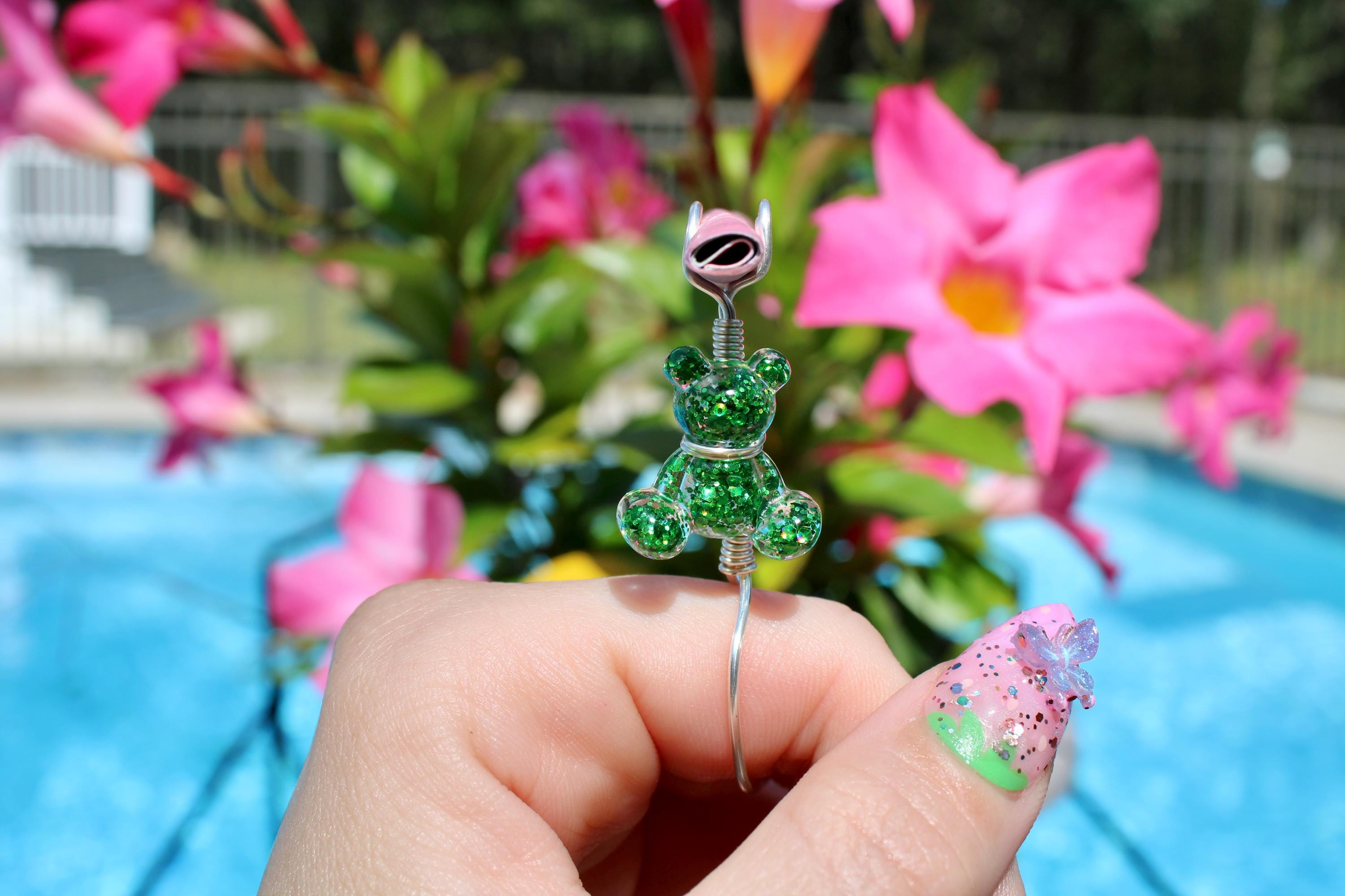 A hand with decorated nails holds a whimsical, green glitter teddy bear smoke ring against a vibrant backdrop of pink flowers and a swimming pool.