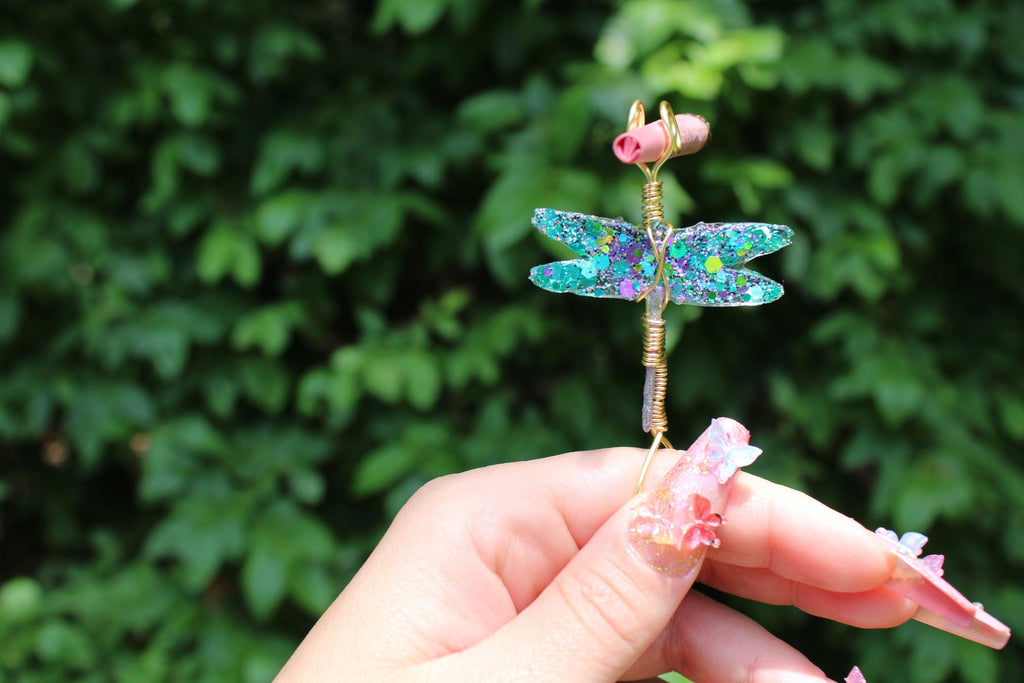 A hand with elaborate floral nail art holds a sparkling, glittery dragonfly smoke ring against a backdrop of green foliage.
