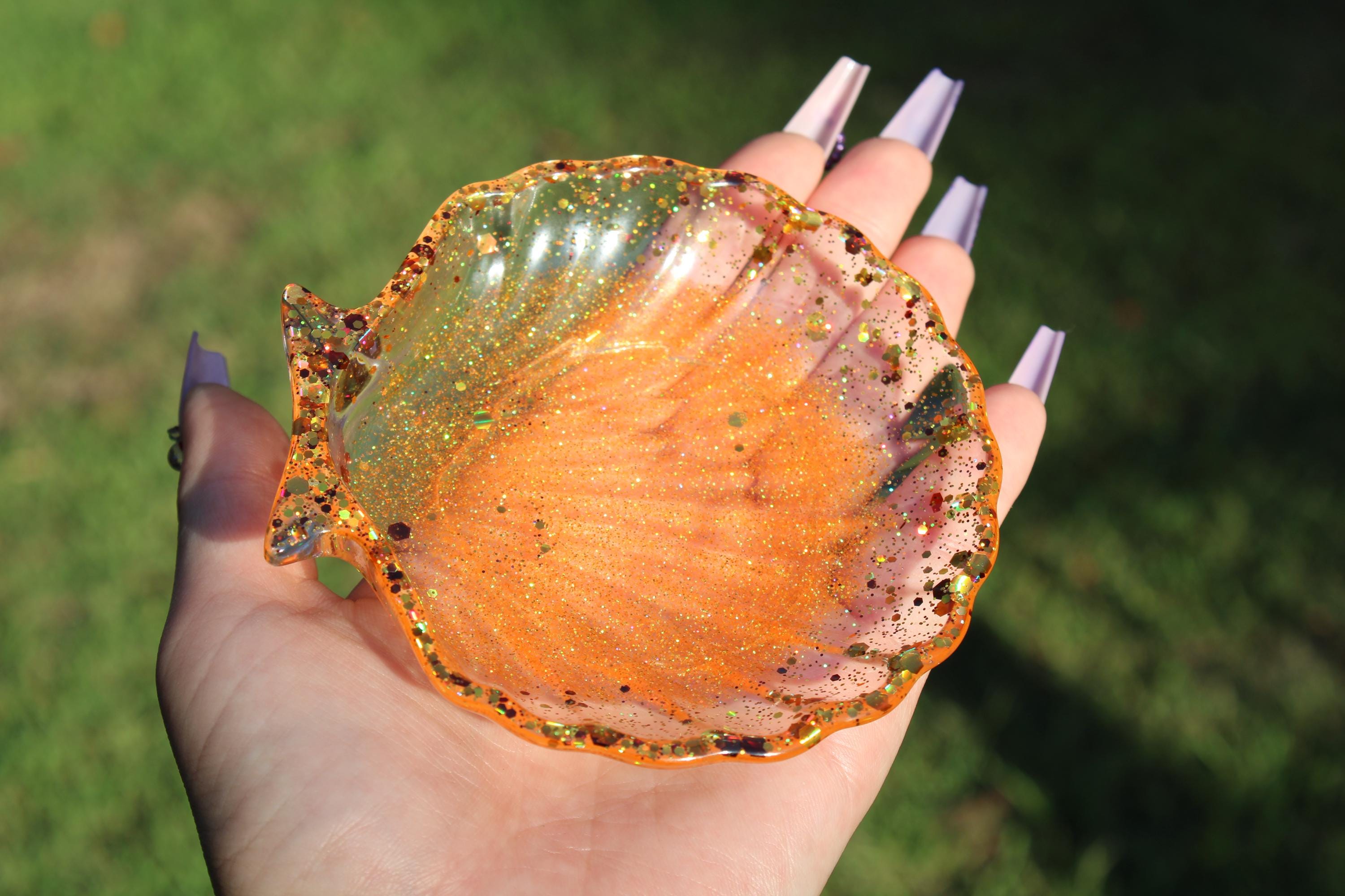 A hand with long, lavender nails holds a shimmery, orange resin seashell trinket dish outdoors.