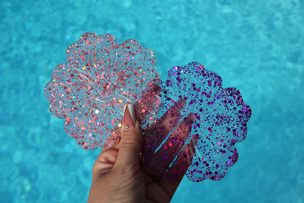 Hand holding two glittery, flower-shaped drink coasters against a blue water background