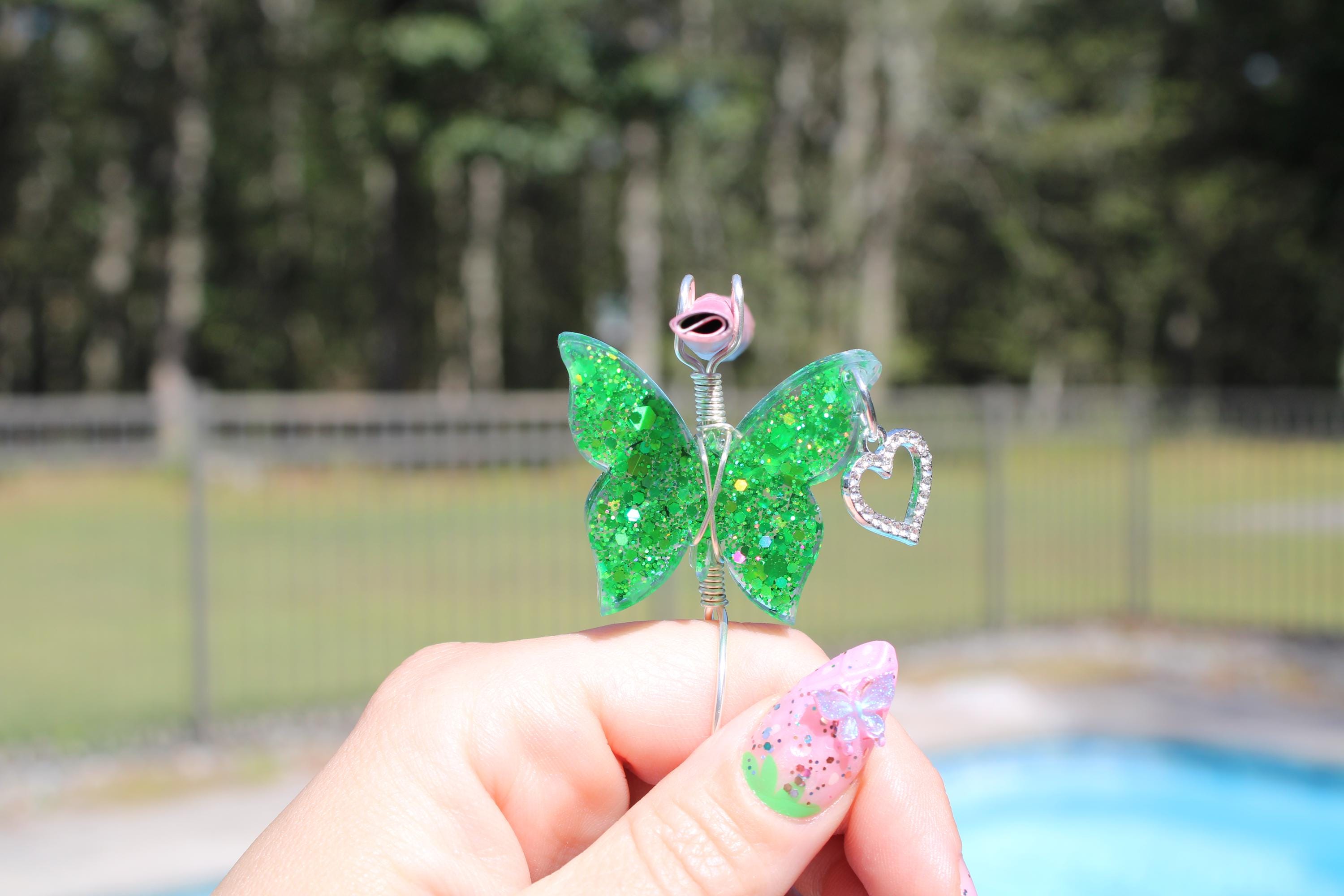 A hand with a decorated nail holds a whimsical green glitter butterfly smoke ring with a heart.