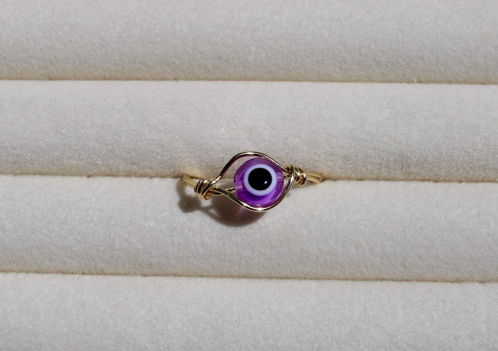 A handcrafted wire ring showcases a vibrant purple evil eye bead against a soft, textured cream surface.