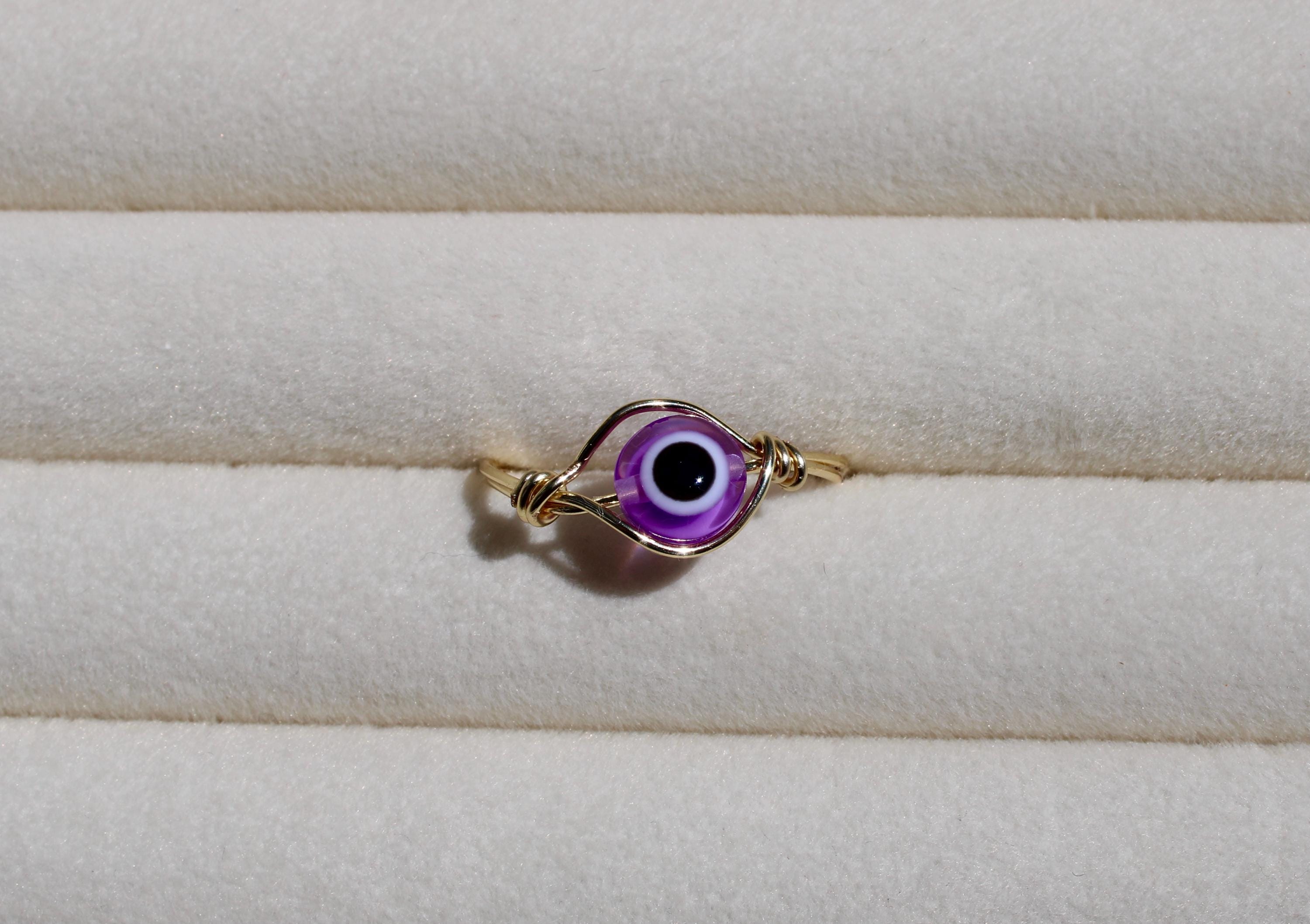 A handcrafted wire ring showcases a vibrant purple evil eye bead against a soft, textured cream surface.