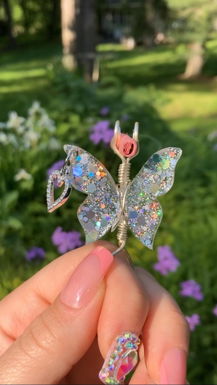 A manicured hand holds a handmade, sparkly butterfly smoke ring with a heart charm in a garden of flowers.