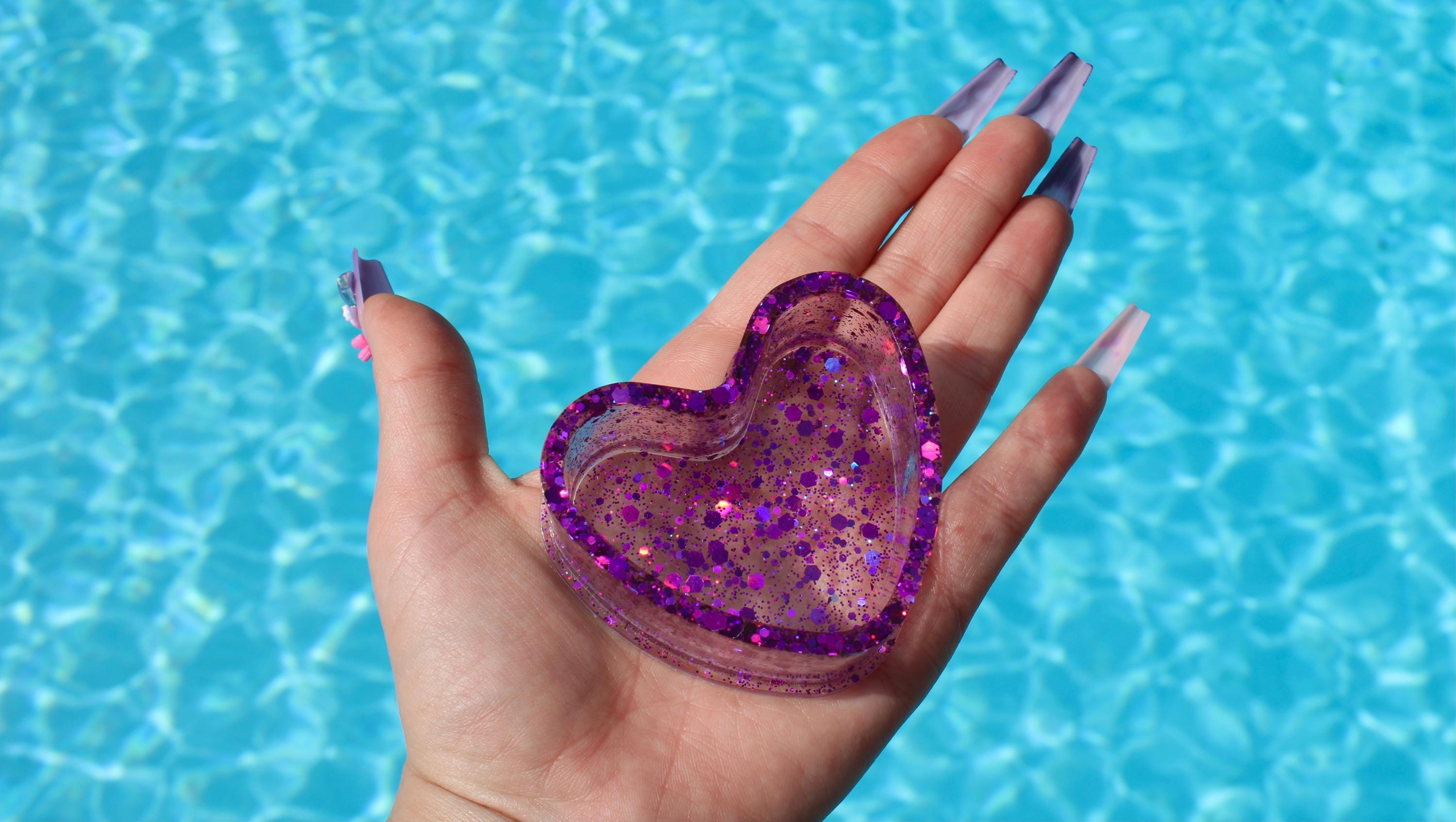 Hand holding a purple glittery heart-shaped ring dish with a pool background