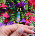 Woman's manicured hand displays a blue and pink aura quartz crystal smoke ring against a floral backdrop.