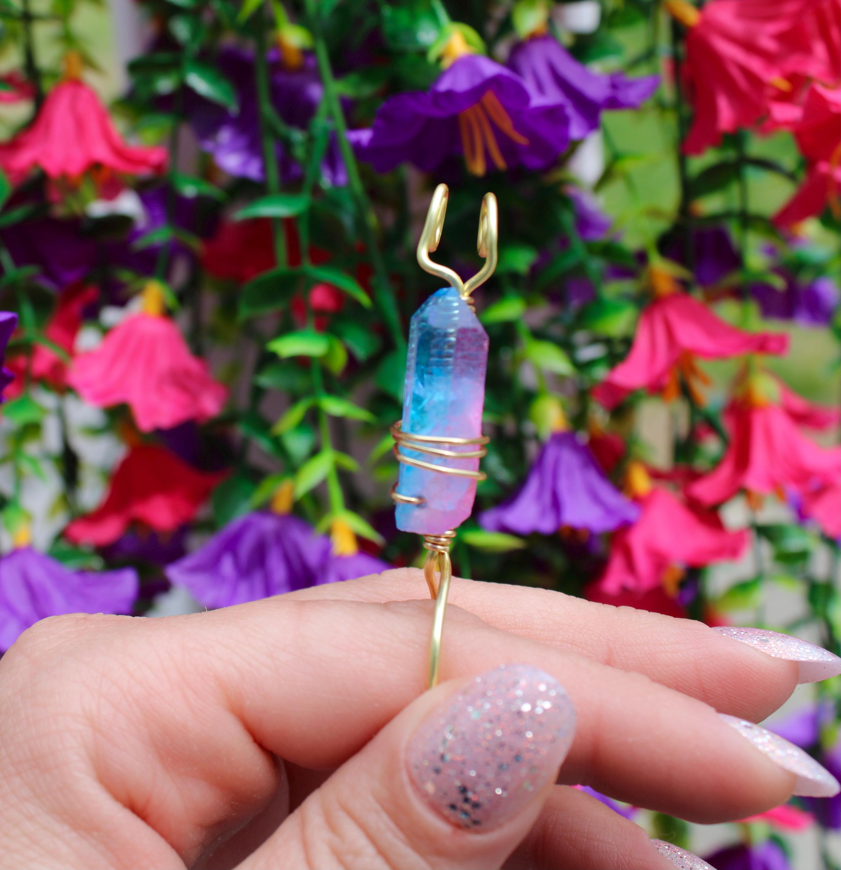 Woman's manicured hand displays a blue and pink aura quartz crystal smoke ring against a floral backdrop.