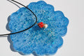 Close-up showcases a whimsical red and white mushroom pendant with gold wire detail, resting on a sparkly blue resin coaster.
