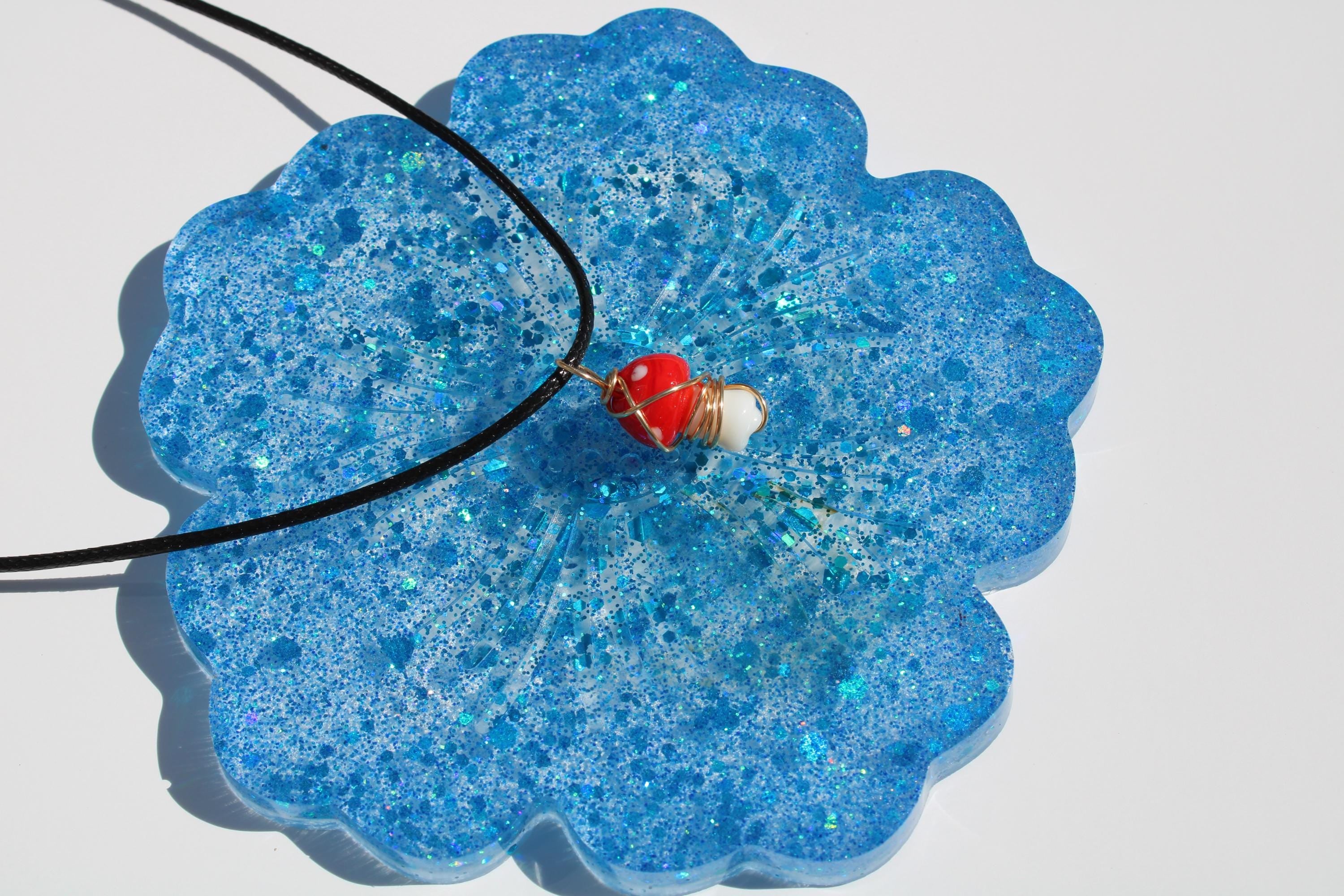 Close-up showcases a whimsical red and white mushroom pendant with gold wire detail, resting on a sparkly blue resin coaster.