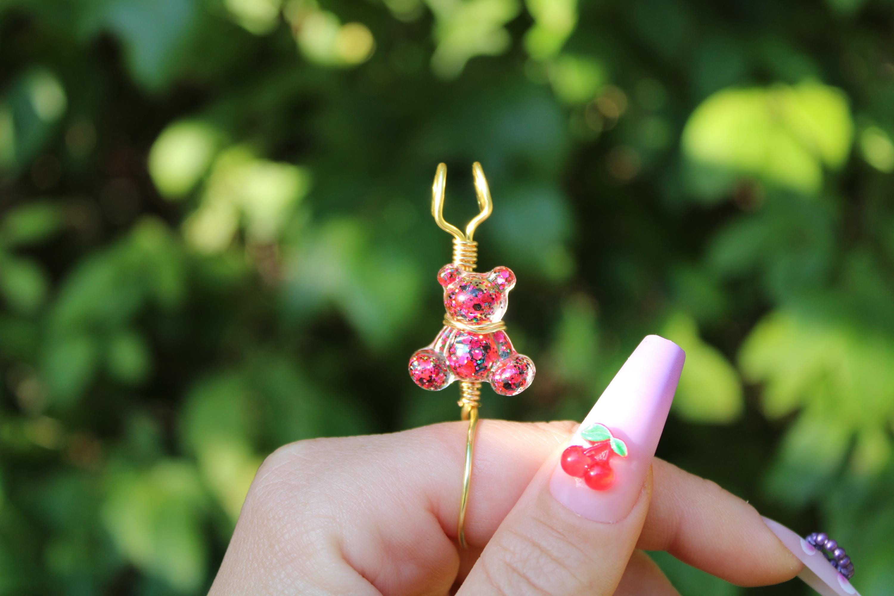 A hand with lavender, cherry-accented nails holds a pink sequin teddy bear smoke ring against a leafy background.