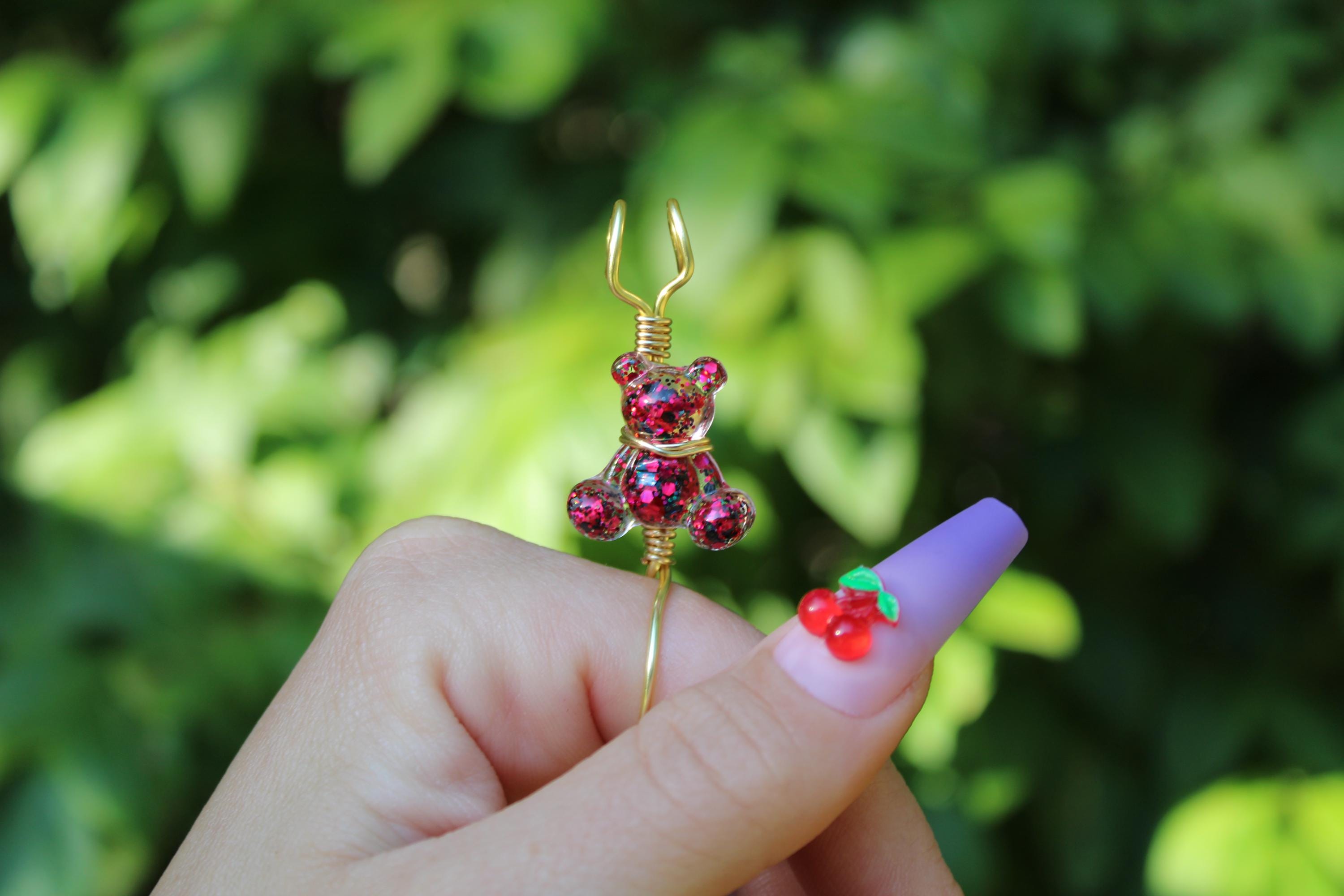 A hand model holds a pink sequin teddy bear smoke ring with a cherry embellishment on a lavender press-on nail.