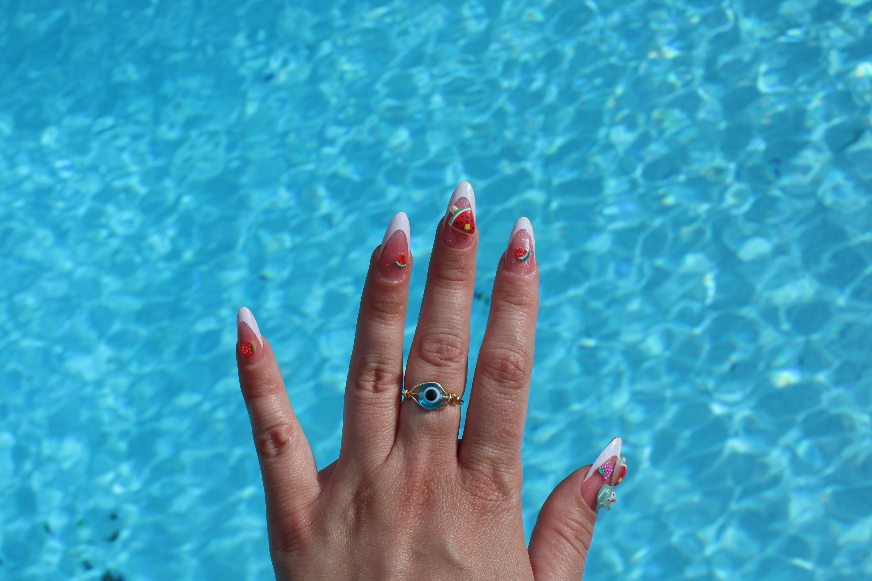 A hand with white-tipped acrylic nails decorated with fruit charms and an evil eye ring poses over shimmering blue pool water.