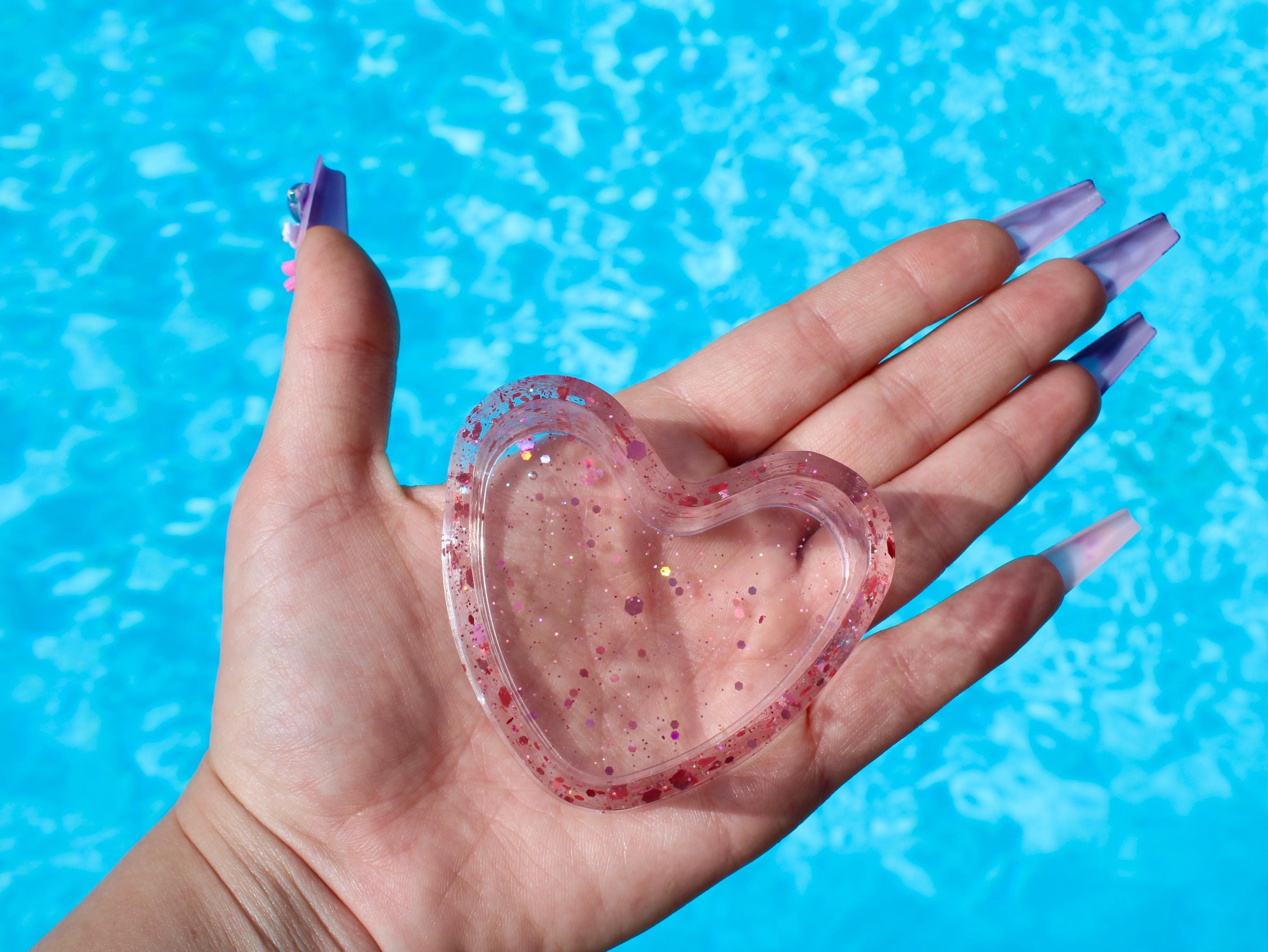 Heart-shaped ring dish with glittery texture held in a hand against a blue water background