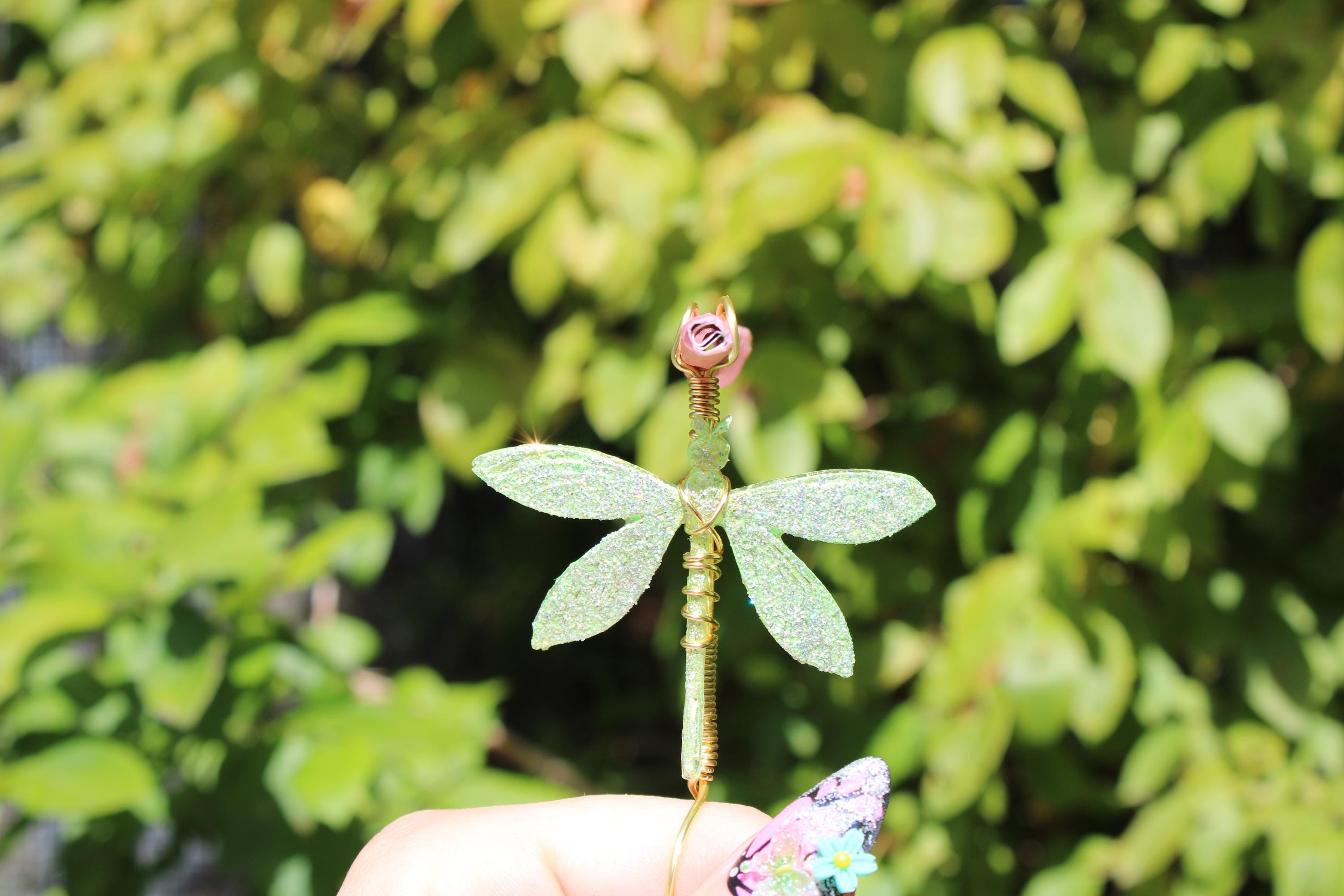A hand holds a sparkly green dragonfly smoke ring against a green bush.