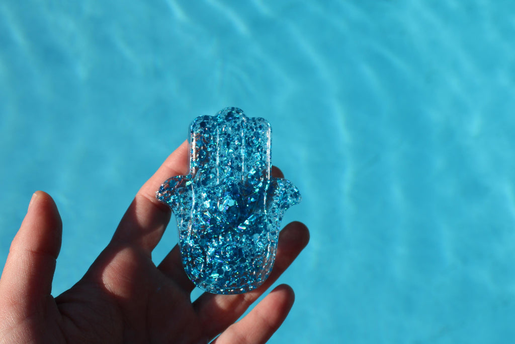 Hand holding a blue glittery hamsa hand against a clear blue water background