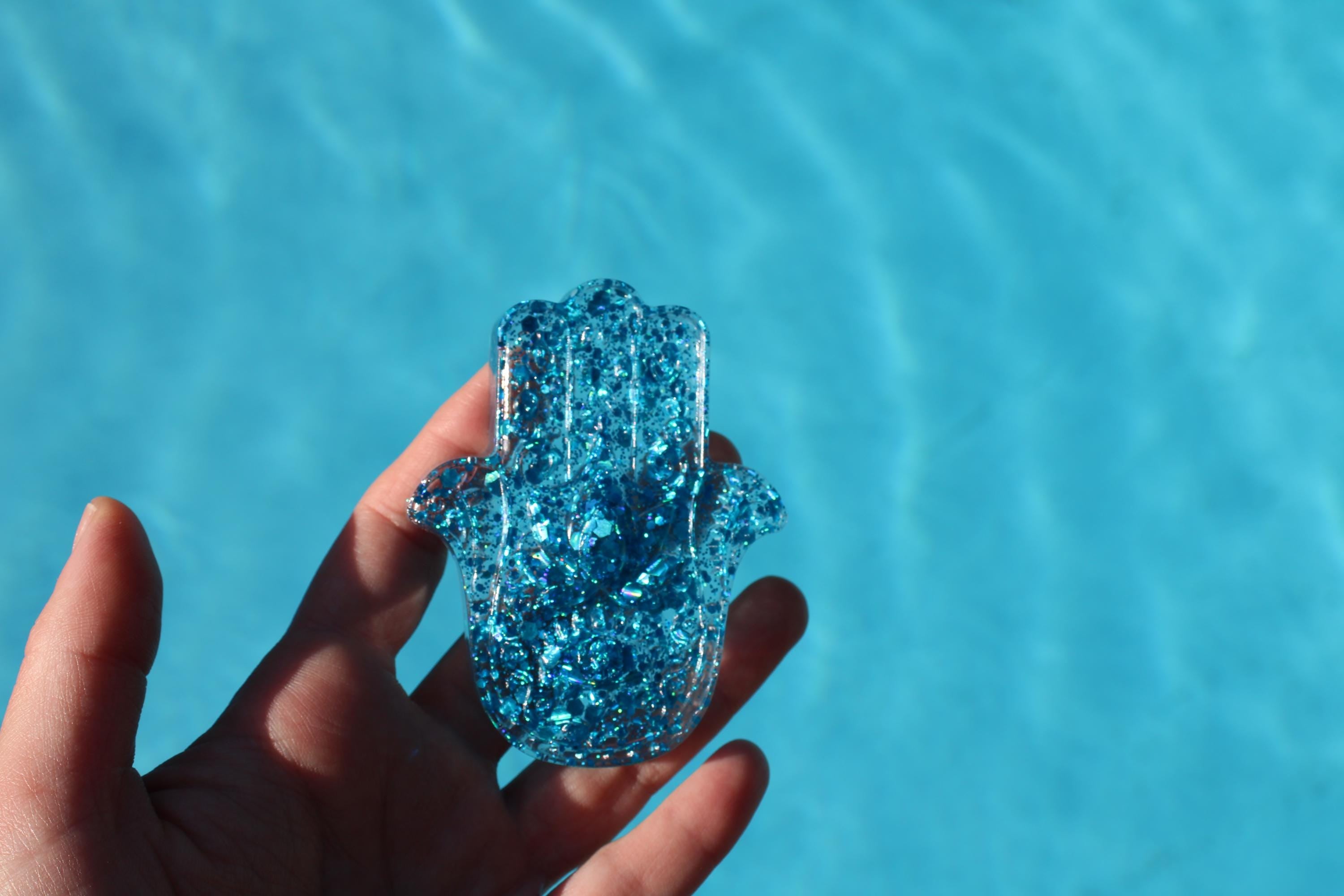 Hand holding a blue glittery hamsa hand against a clear blue water background