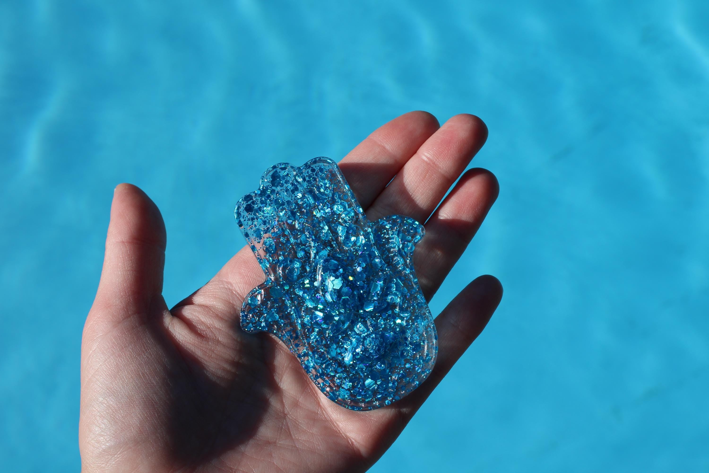 Hand holding a blue glittery hamsa hand against a blue water background