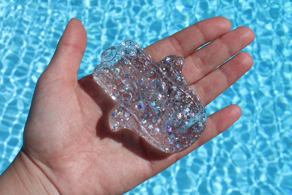 A hand holds a sparkly, clear Hamsa Hand against the background of a shimmering blue swimming pool.