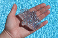 A hand holds a sparkly, clear Hamsa Hand against the background of a shimmering blue swimming pool.