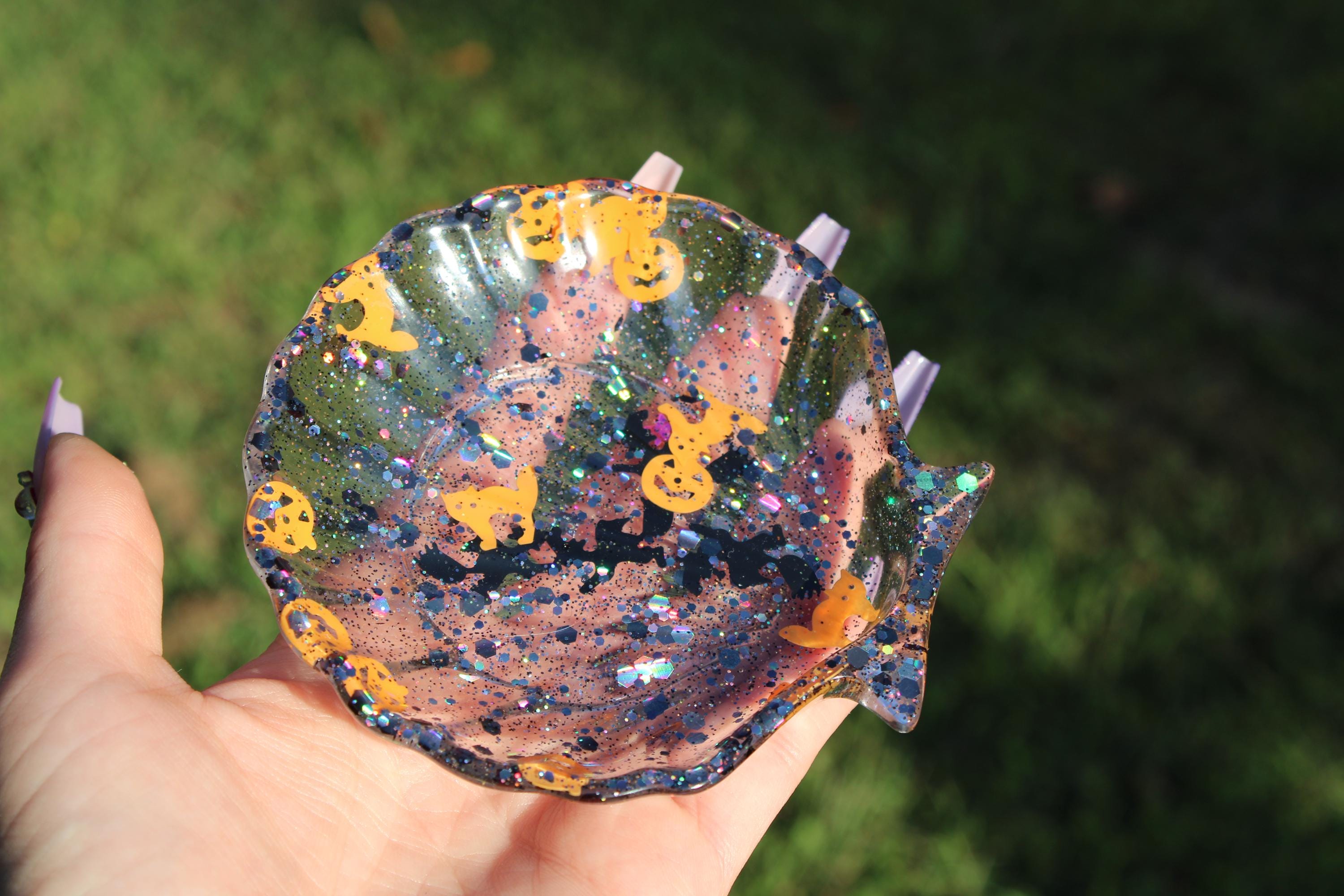 Hand holds a sparkling, handmade resin dish featuring Halloween-themed shapes like jack-o'-lanterns and black cats.