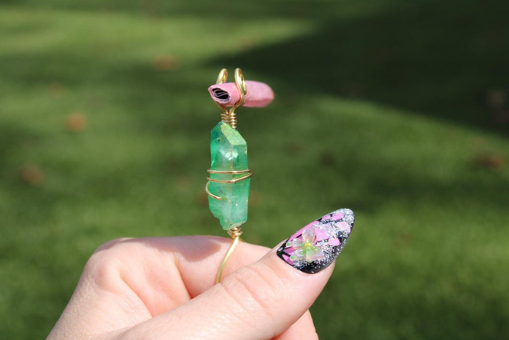 A hand with a glittery floral nail design holds a green crystal smoke ring wrapped with copper gold wire.