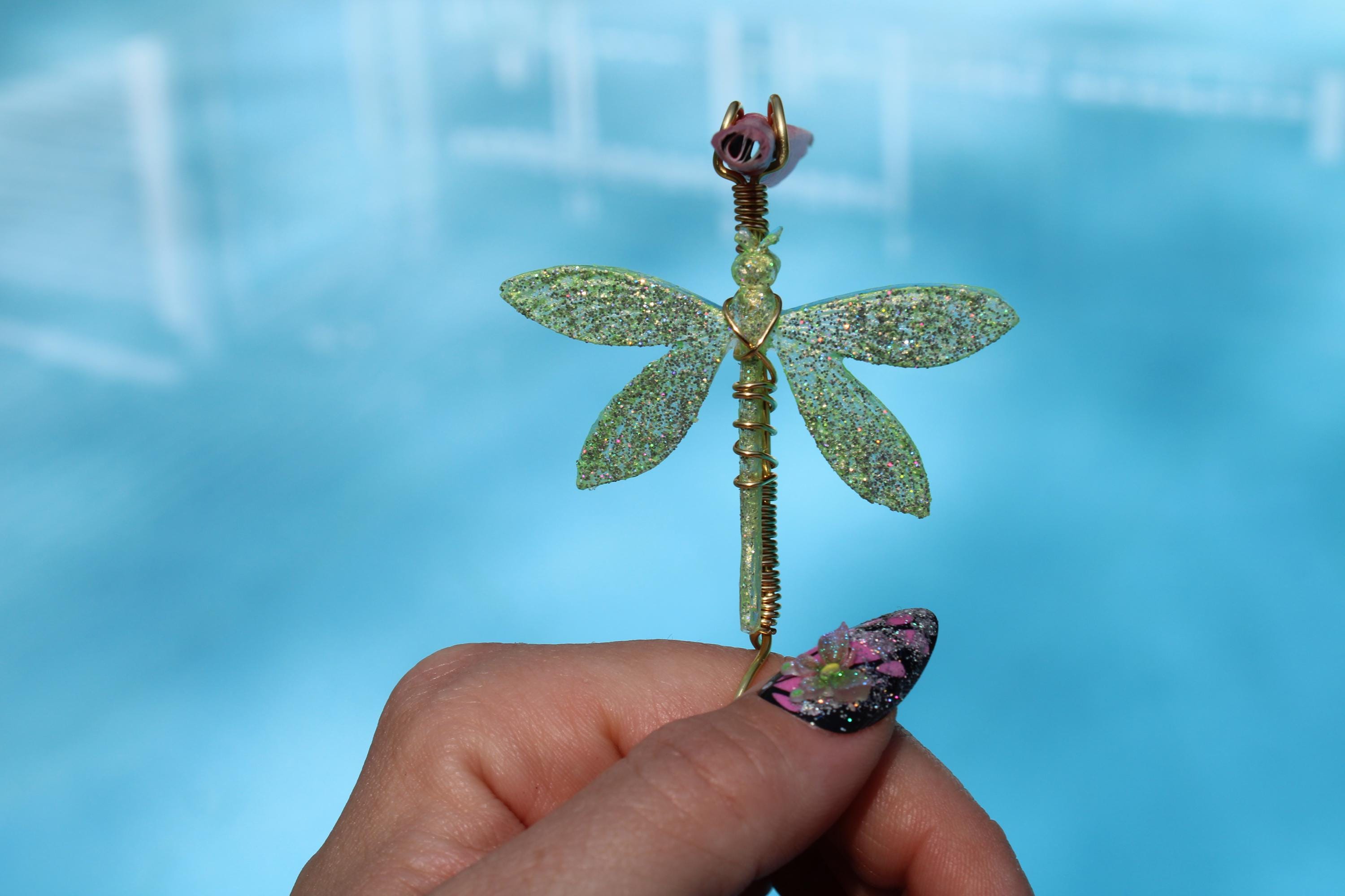 A hand with a floral nail design holds a whimsical, glittery, wire-wrapped dragonfly smoking accessory craft against a blue background.
