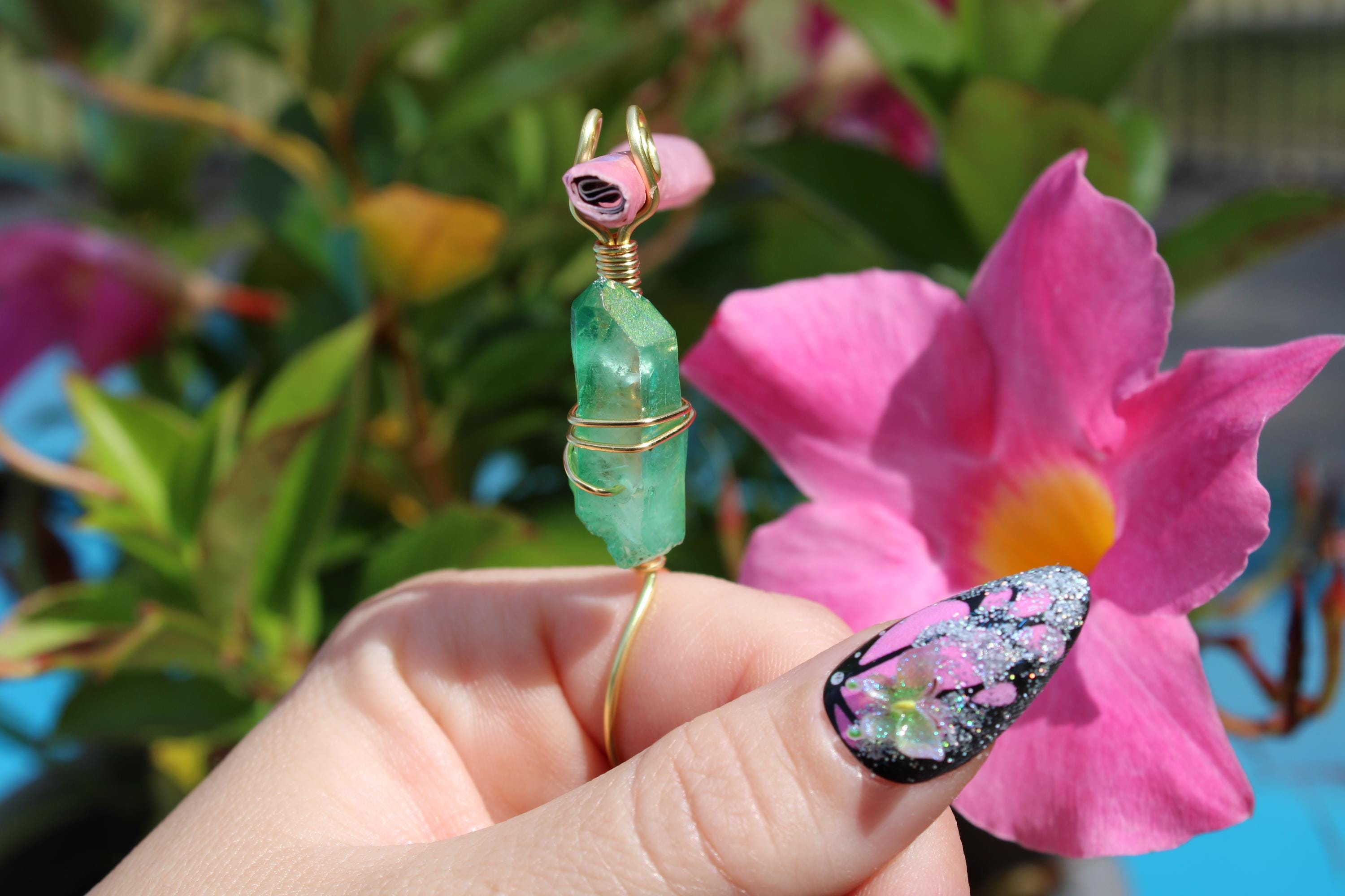 A hand with a glittery floral-art manicure holds a wire-wrapped green crystal smoke ring near a vibrant pink flower.
