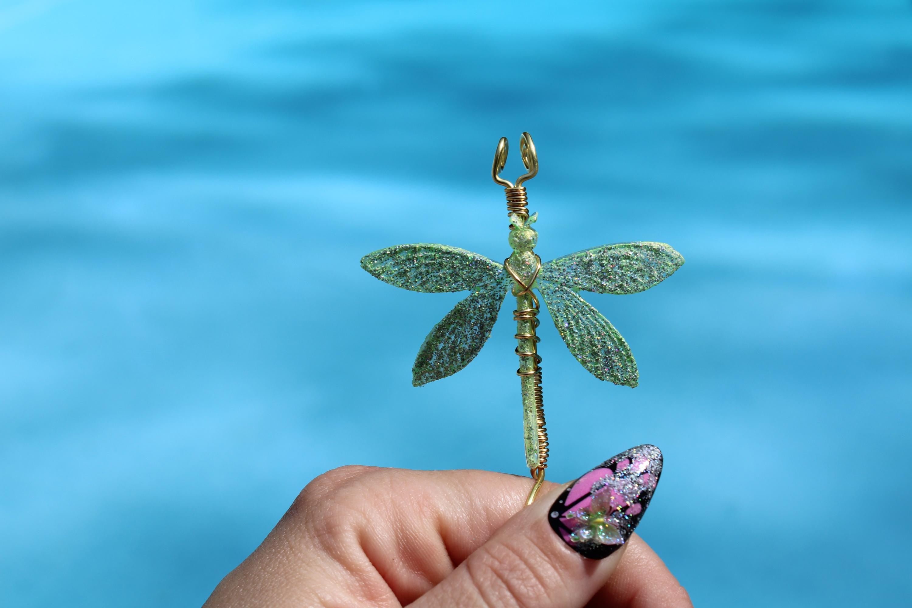 A hand with glittery nail art holds a whimsical, handmade gold wire and green glitter dragonfly smoke ring against a blurry blue background.