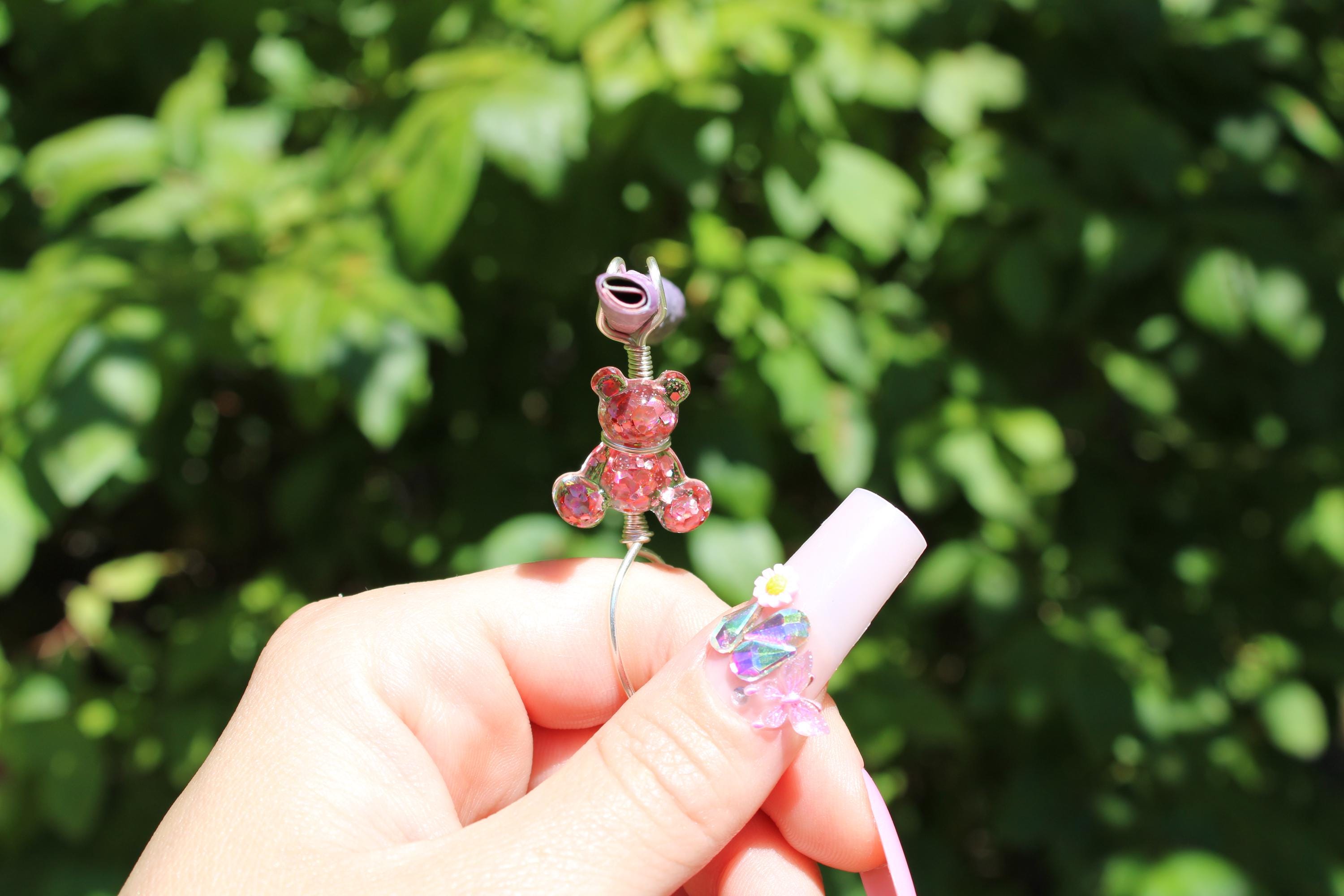 A hand with long pink acrylic nails decorated with butterflies holds a handmade wire smoke ring featuring a sparkling teddy bear and purple flower embellishment against a leafy background.