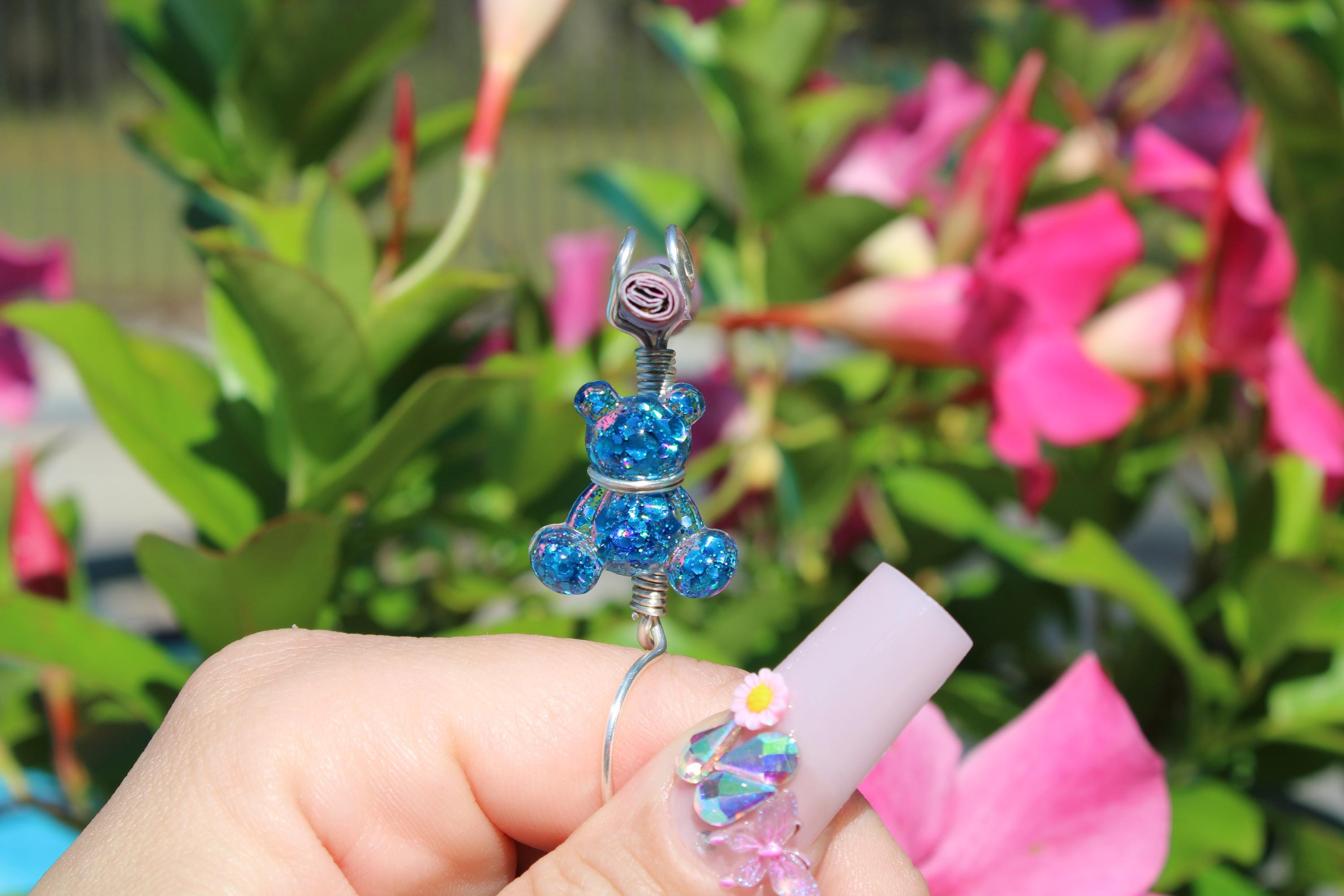 Woman displays a handcrafted silver ring featuring a blue glitter teddy bear smoke ring complemented by decorative nail art, set against a vibrant floral background.