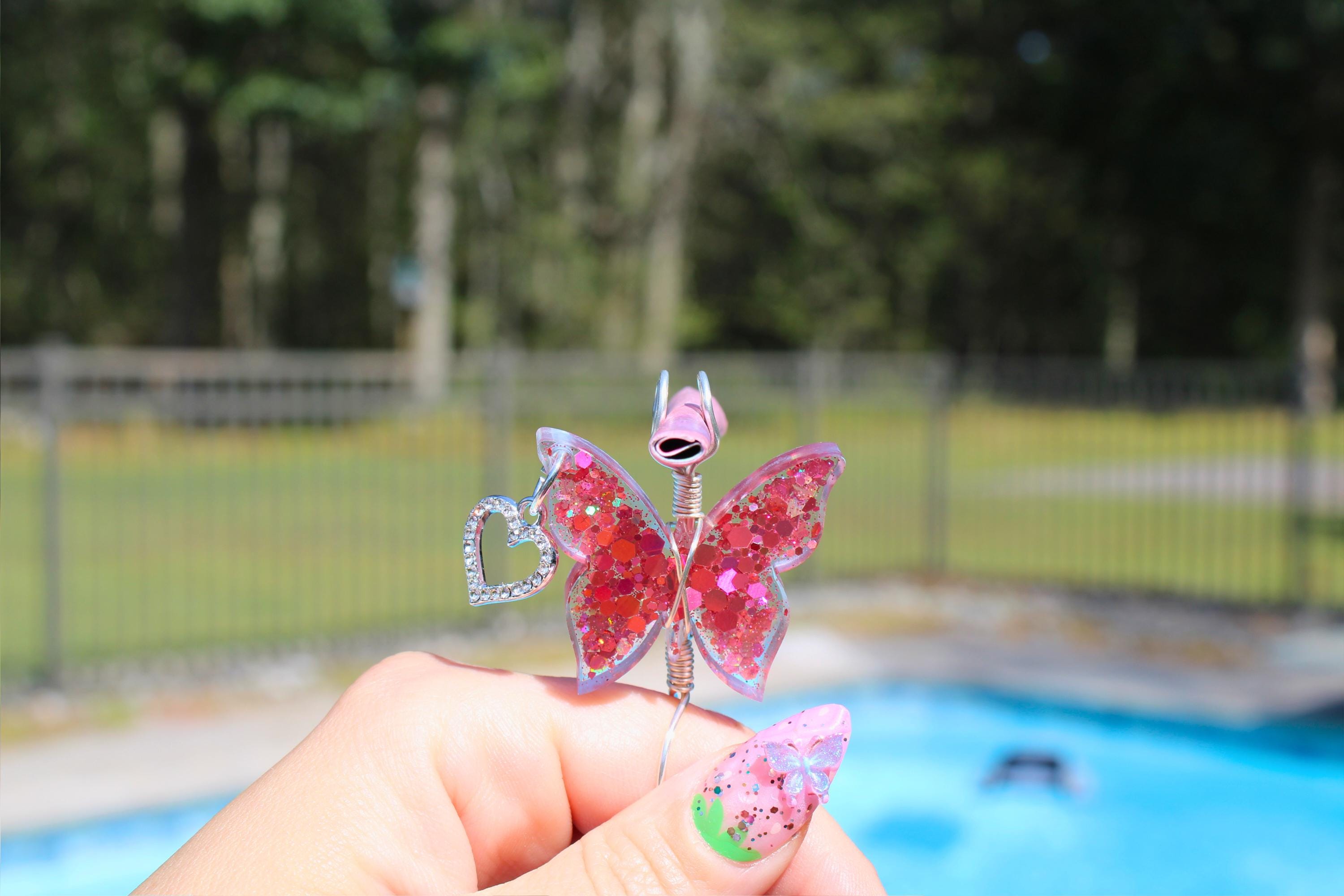 Woman holds a pink butterfly smoke ring with a dangling heart charm in front of a pool.