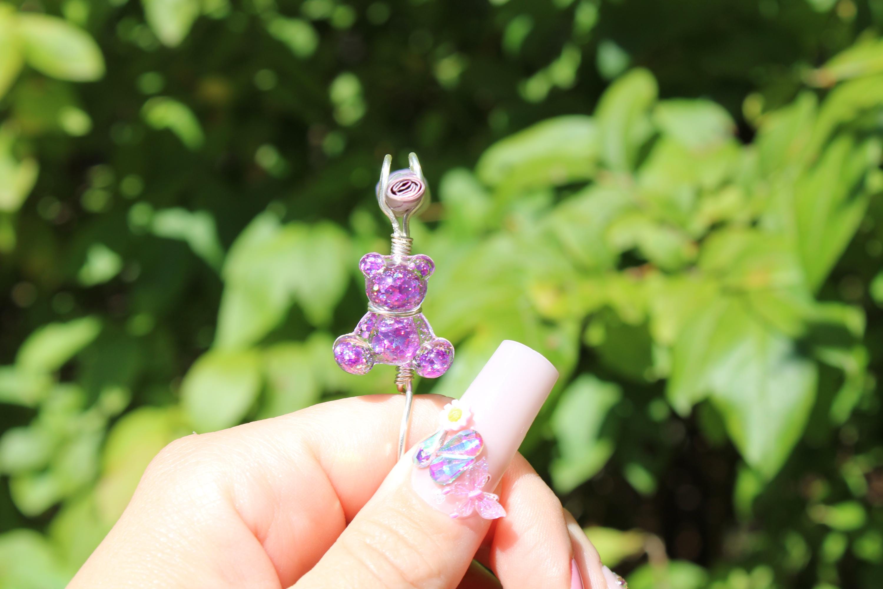 A manicured hand holds a handmade purple glitter teddy bear smoke ring against a green foliage background.