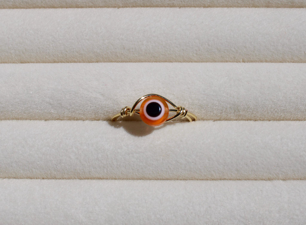 A wire ring featuring an orange and black evil eye bead sits on a ribbed, off-white surface.