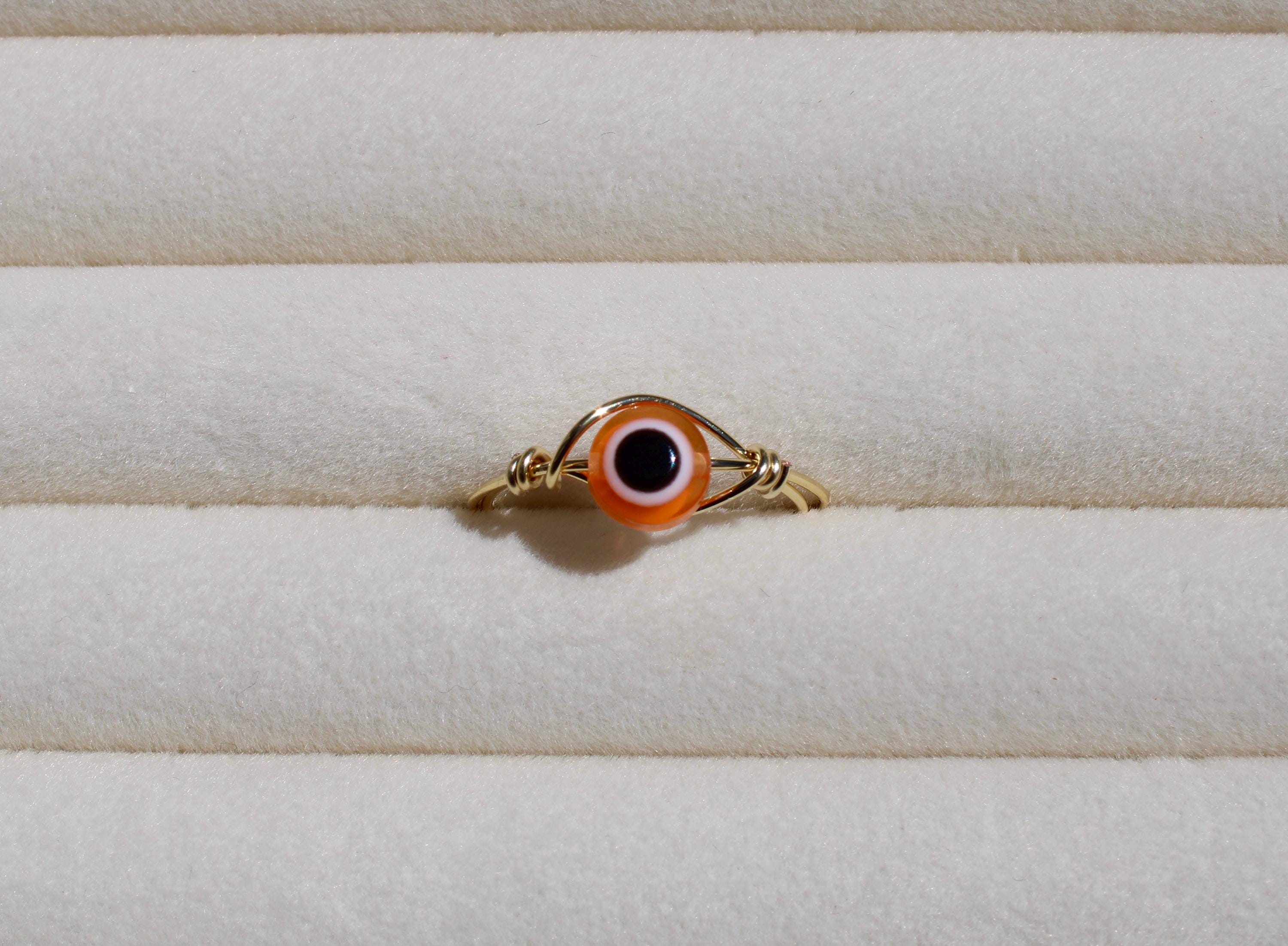 A wire ring featuring an orange and black evil eye bead sits on a ribbed, off-white surface.