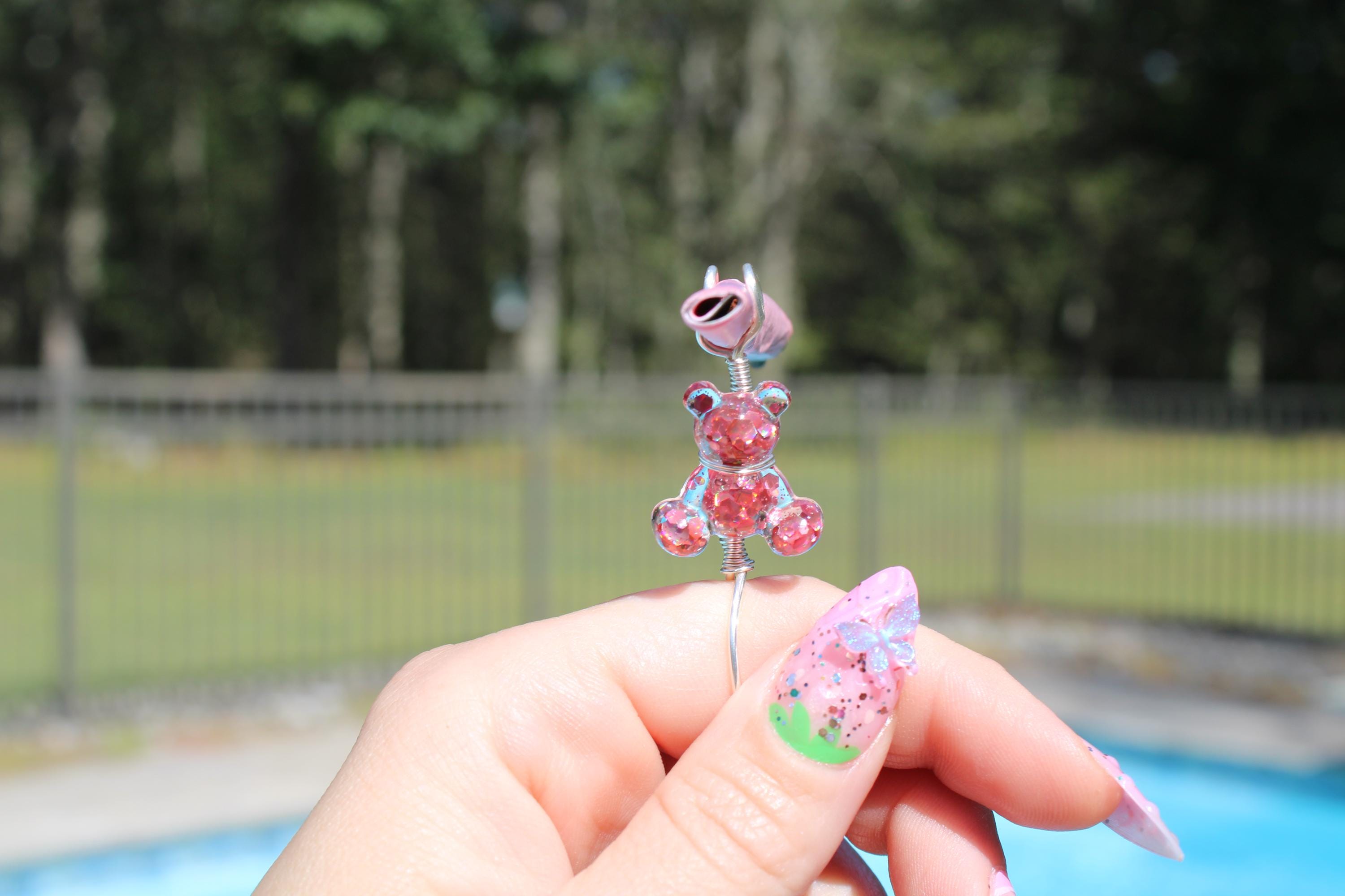 A hand with elaborately decorated pink nails displays a handmade jewelry piece featuring a glittery pink teddy bear smoke ring.